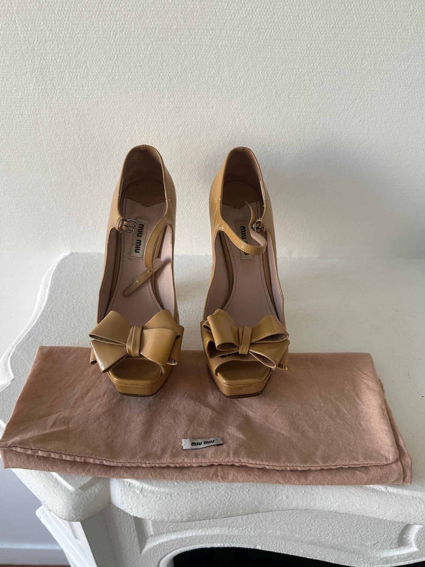 A pair of beige high-heeled shoes with bows on the toe and heel, placed on a white surface.