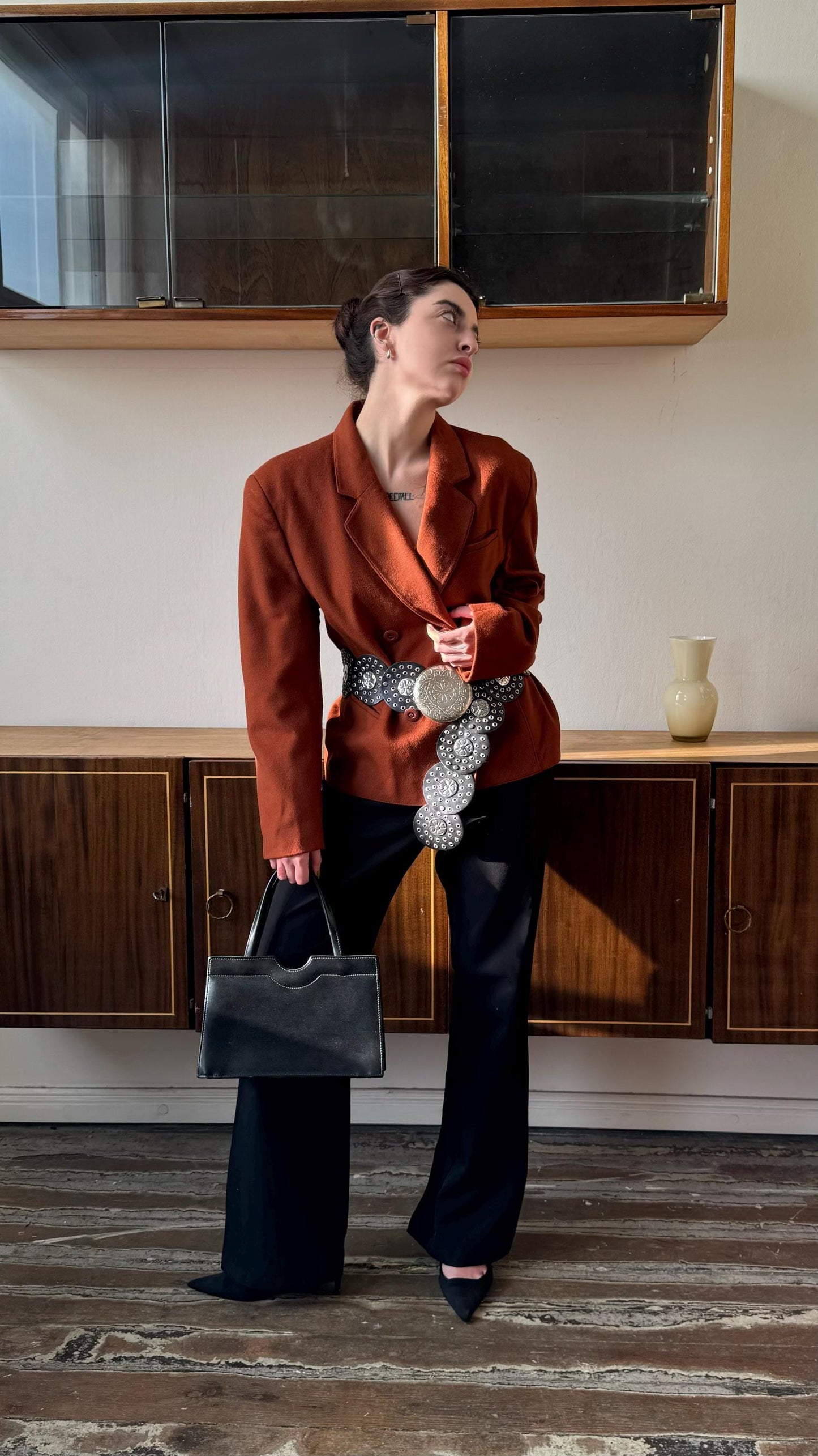 A woman in a red blazer and black pants stands in a room, holding a black handbag and looking up.