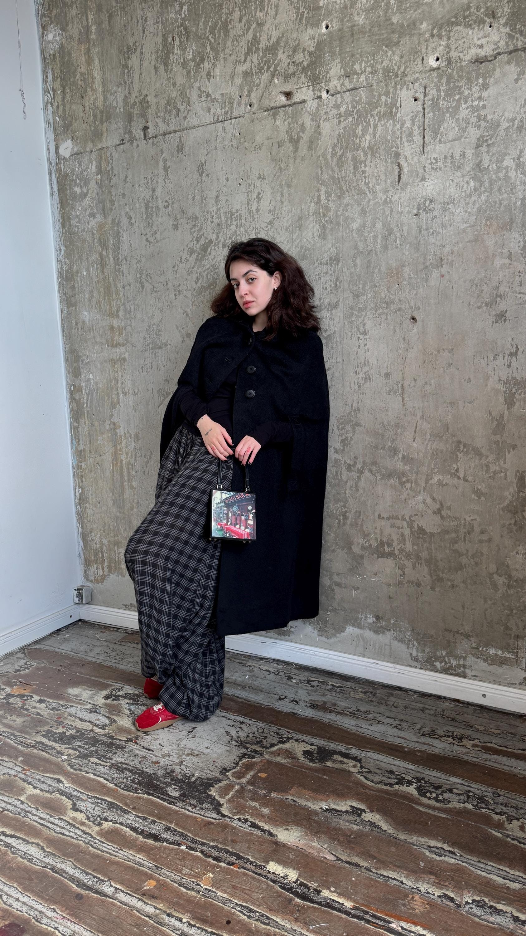 A woman in a black coat and plaid skirt stands against a concrete wall, holding a bag with a picture on it.