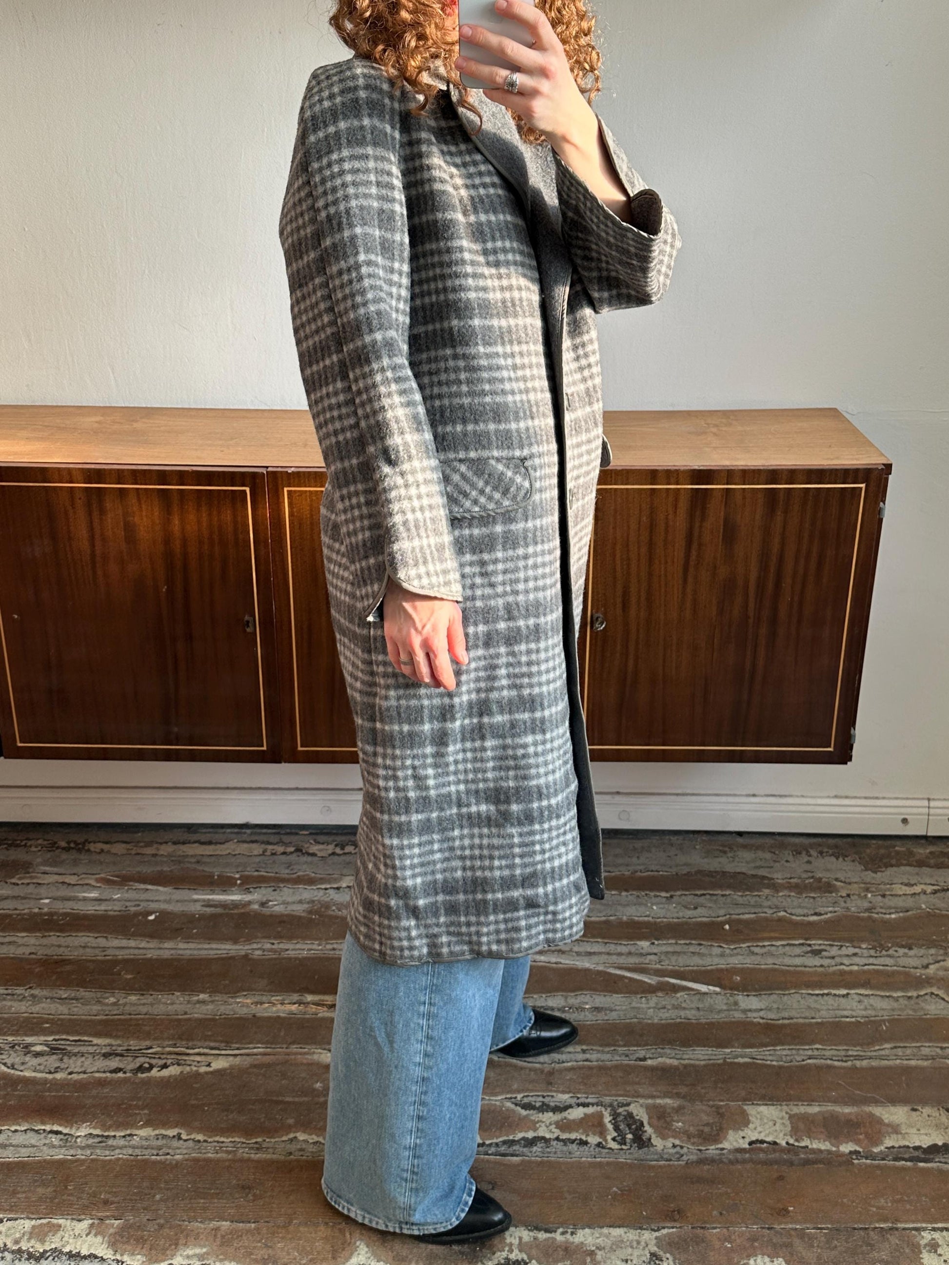 A person is taking a selfie with a plaid coat and jeans, standing in front of a wooden cabinet.