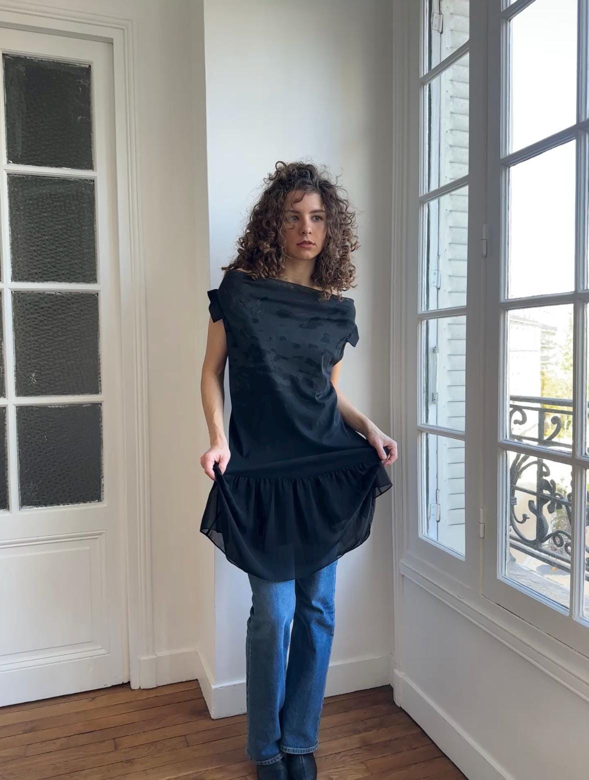 A woman with curly hair stands in a room, wearing a black dress and blue jeans.