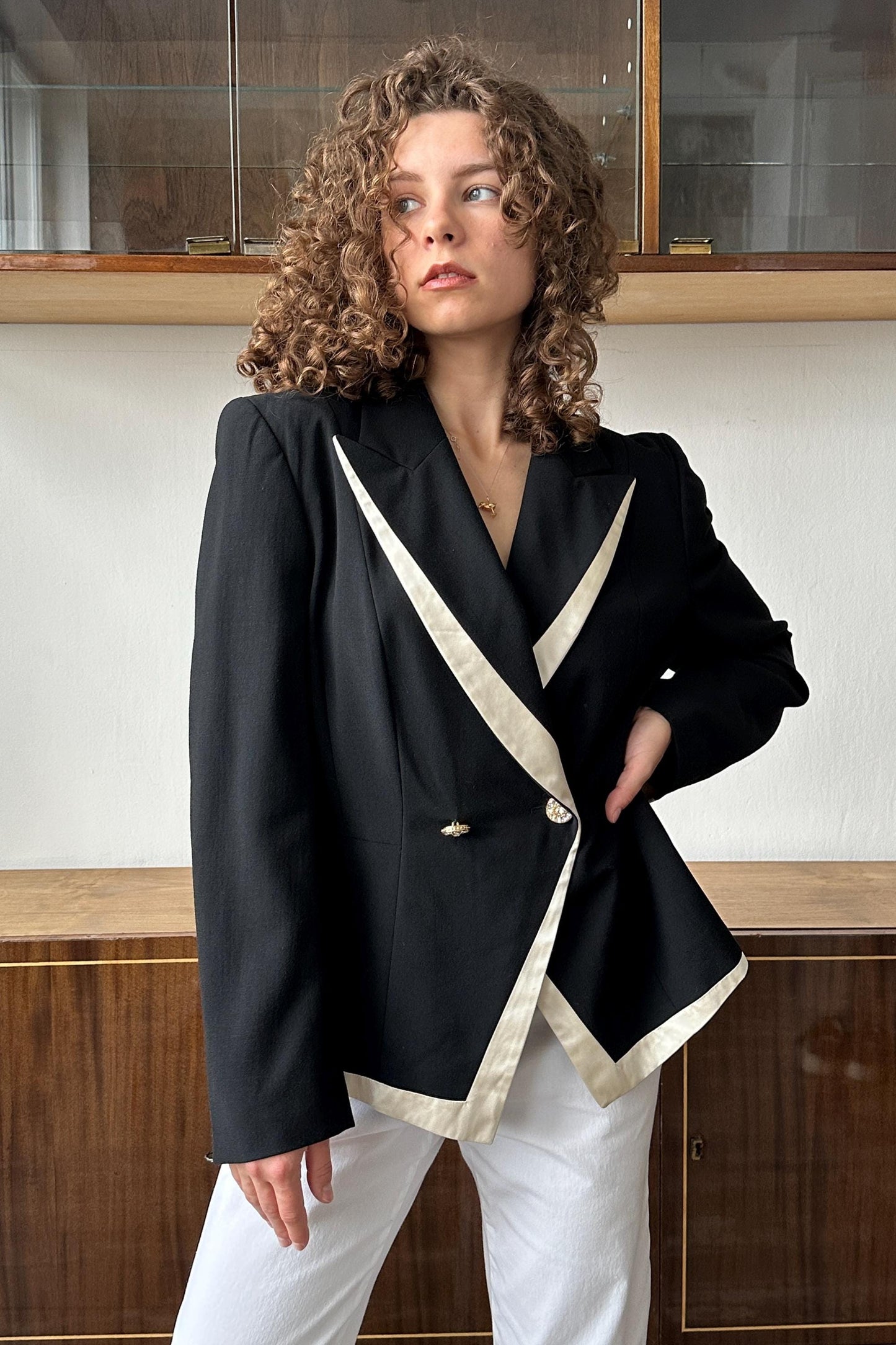 A woman with curly hair wearing a black and white blazer stands in front of a wooden cabinet.