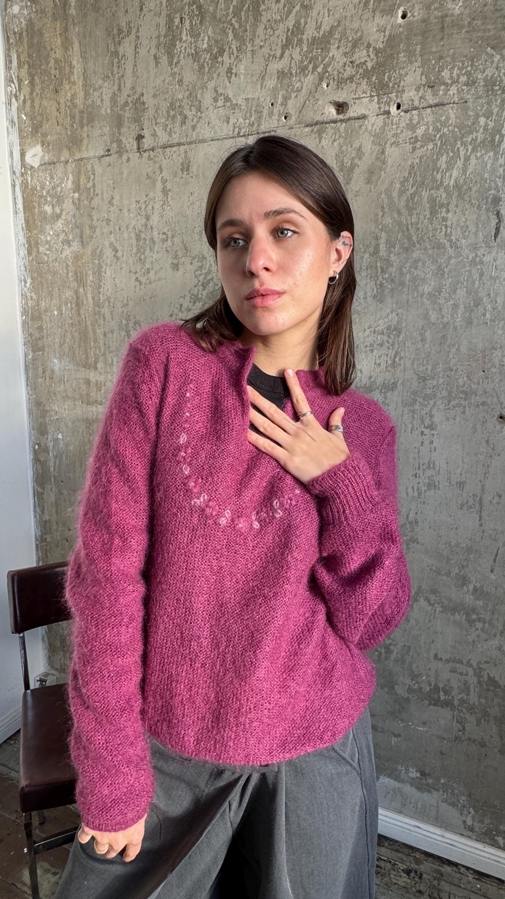 A young woman wearing a pink sweater stands in front of a concrete wall, looking directly at the camera with a thoughtful expression.