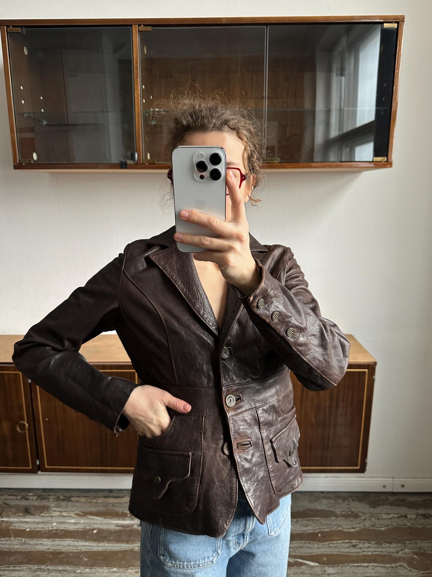 A person is taking a selfie with a smartphone, wearing a brown leather jacket and jeans, in front of a wooden cabinet.