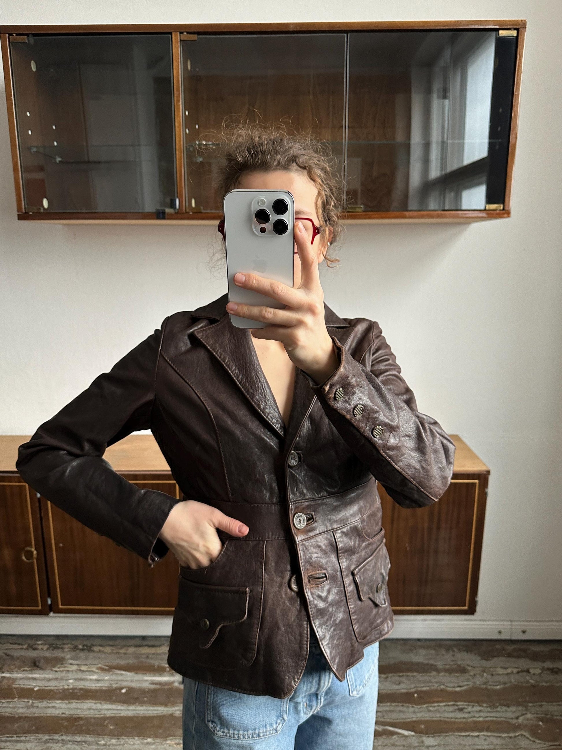 A person is taking a selfie with a smartphone, wearing a brown leather jacket and jeans, in front of a wooden cabinet.