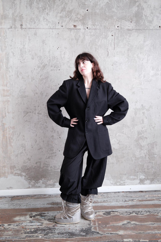 A person wearing a black suit stands in a room with a concrete wall, their arms crossed and hands on their hips.