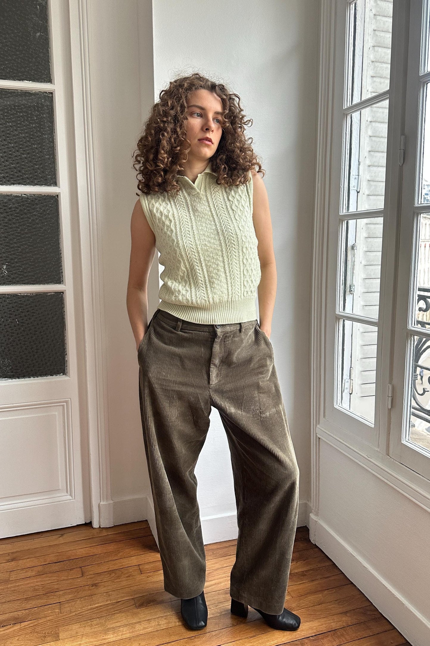 A woman with curly hair stands in a room, wearing a white sweater vest and brown pants.