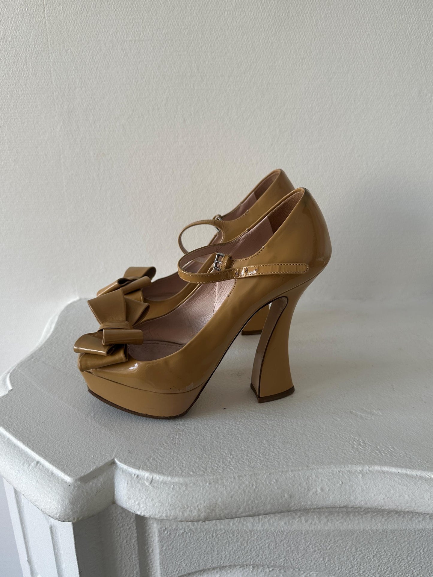A pair of tan high-heeled shoes with bows on the toe and heel, placed on a white surface.