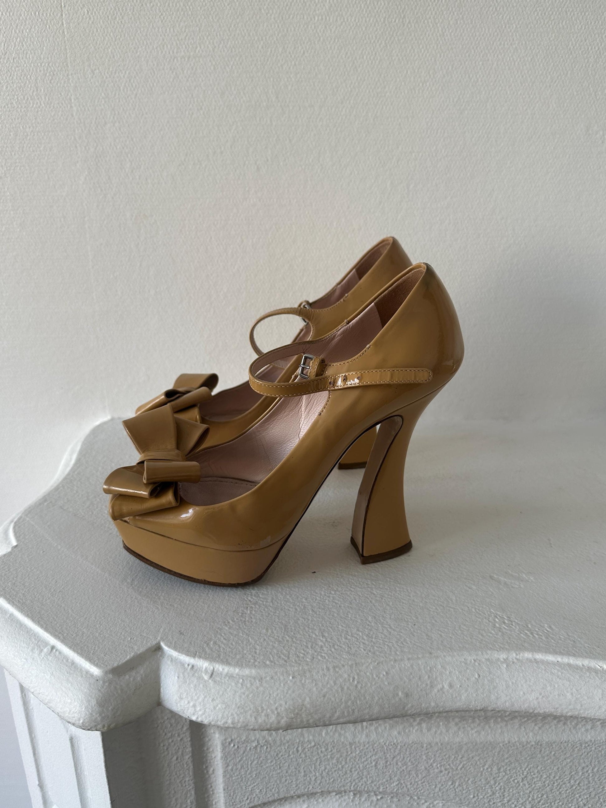A pair of tan high-heeled shoes with bows on the toe and heel, placed on a white surface.