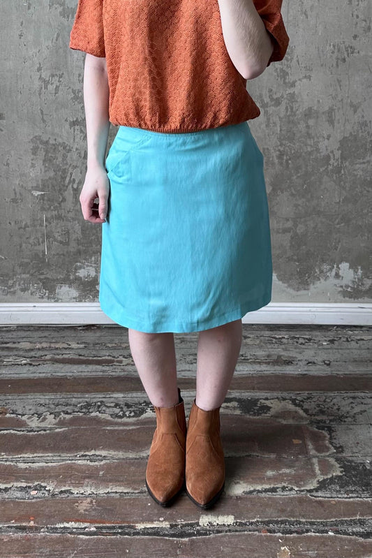 A person wearing a red top, a light blue skirt, and brown boots stands in front of a concrete wall.