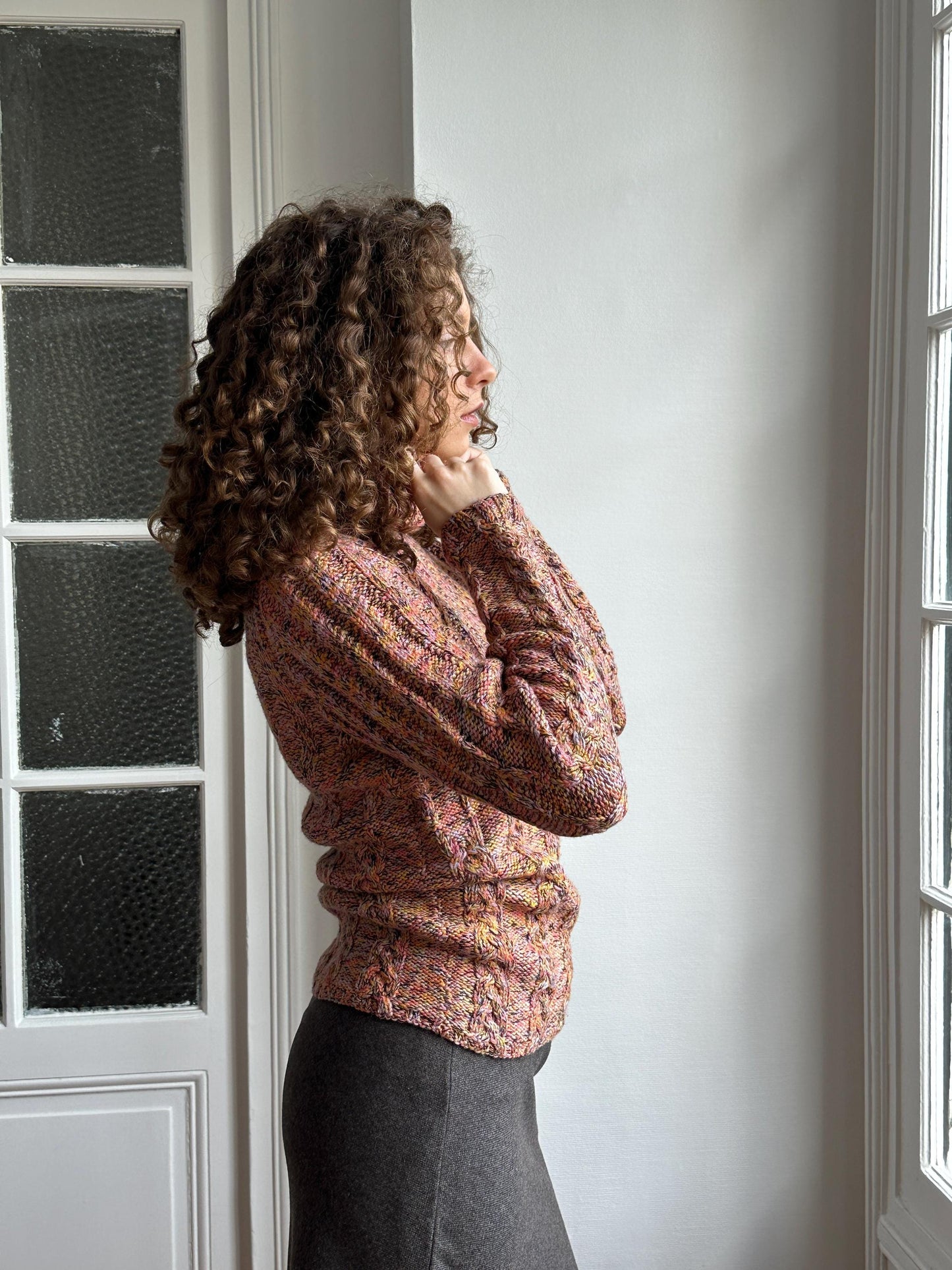 A woman with curly hair is shown in a relaxed pose, wearing a patterned sweater, in an indoor setting with a window in the background.