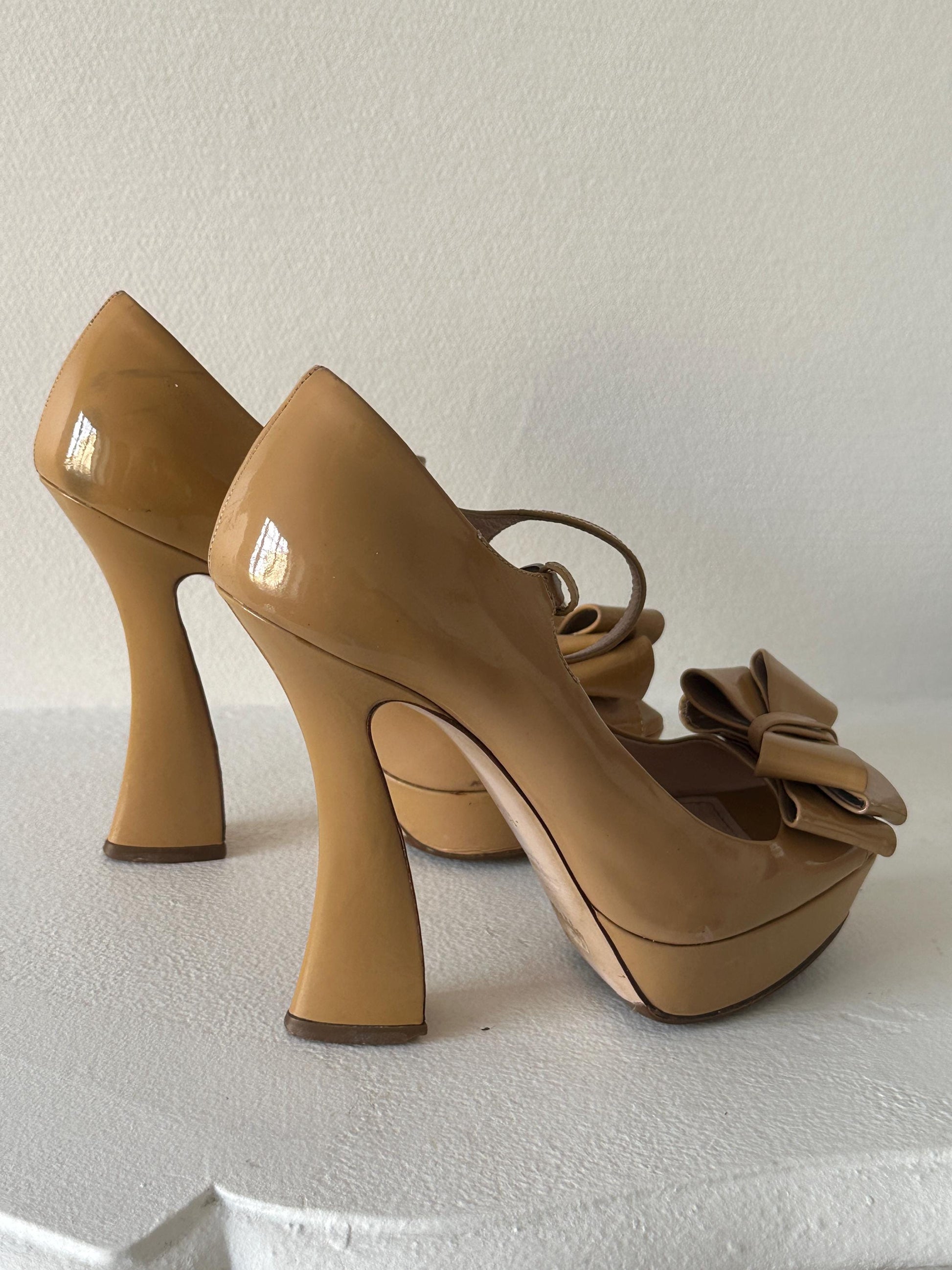 A pair of tan high-heeled shoes with bows on the toe, placed on a white surface.