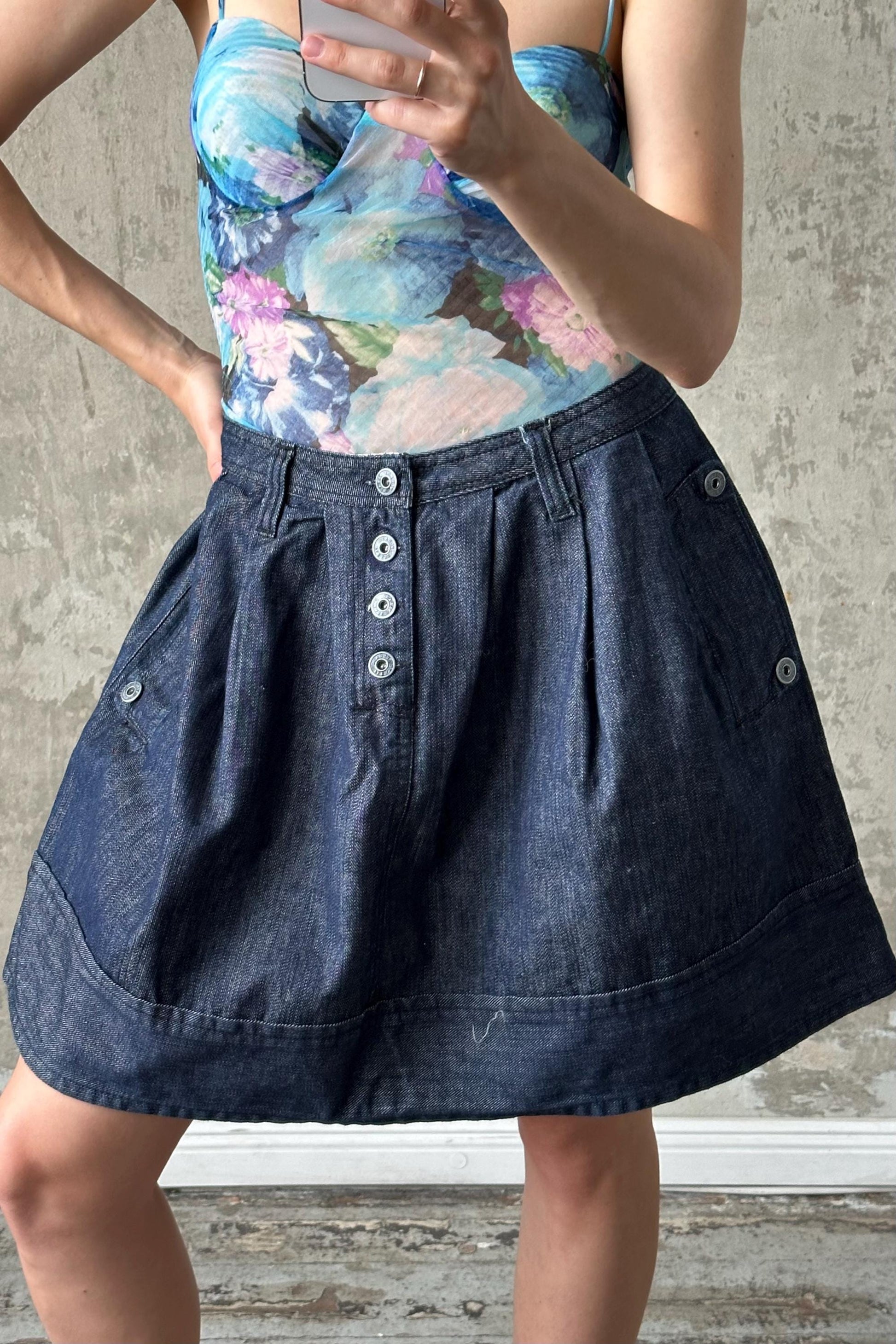A woman wearing a blue denim skirt and a floral top is holding a phone and appears to be taking a photo or recording a video.