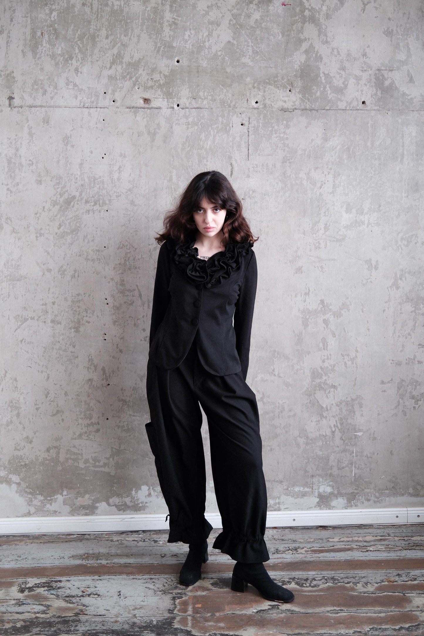 A woman in a black outfit stands in a room with a concrete wall and wooden floor.
