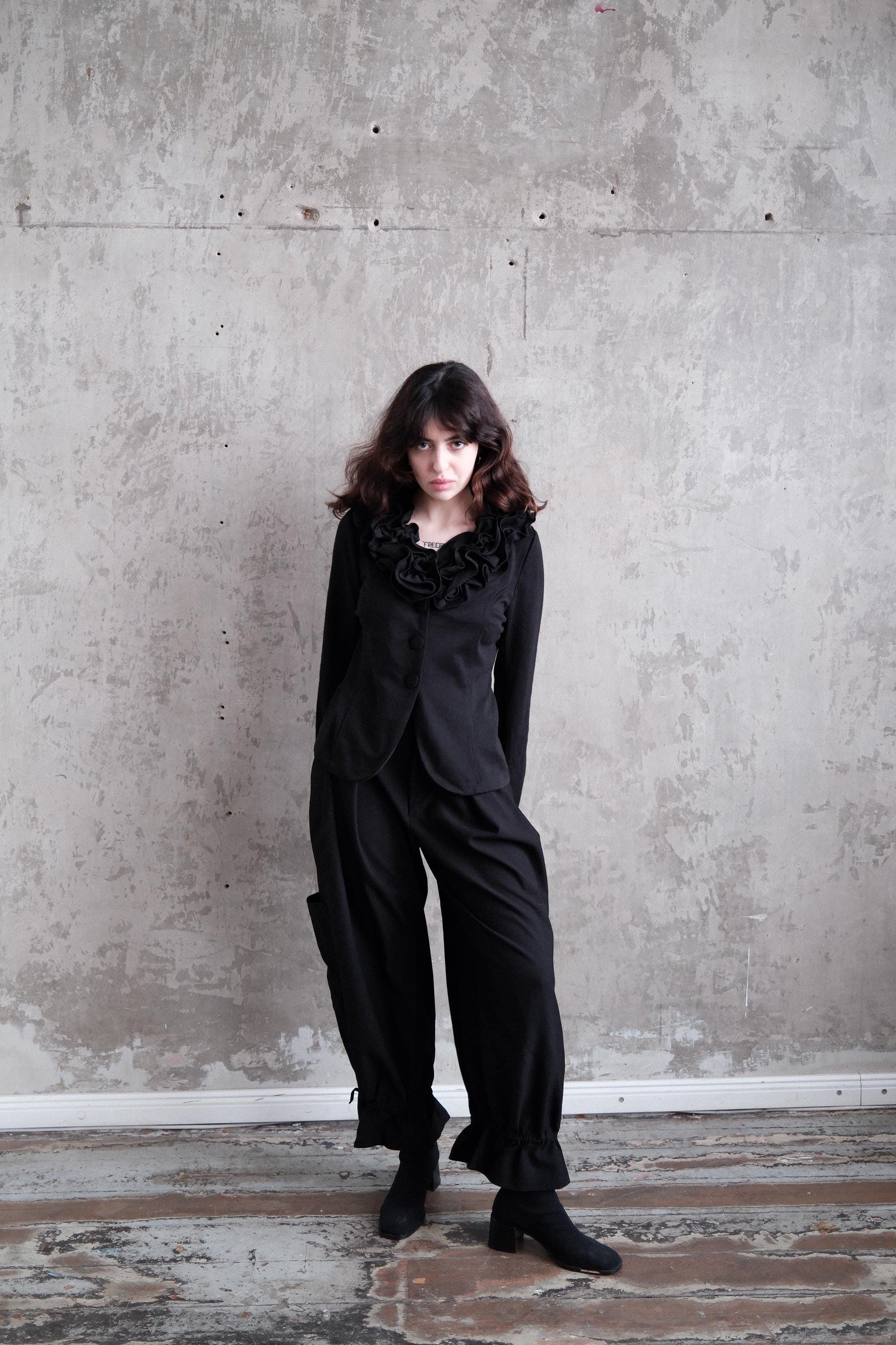 A woman in a black outfit stands in a room with a concrete wall and wooden floor.