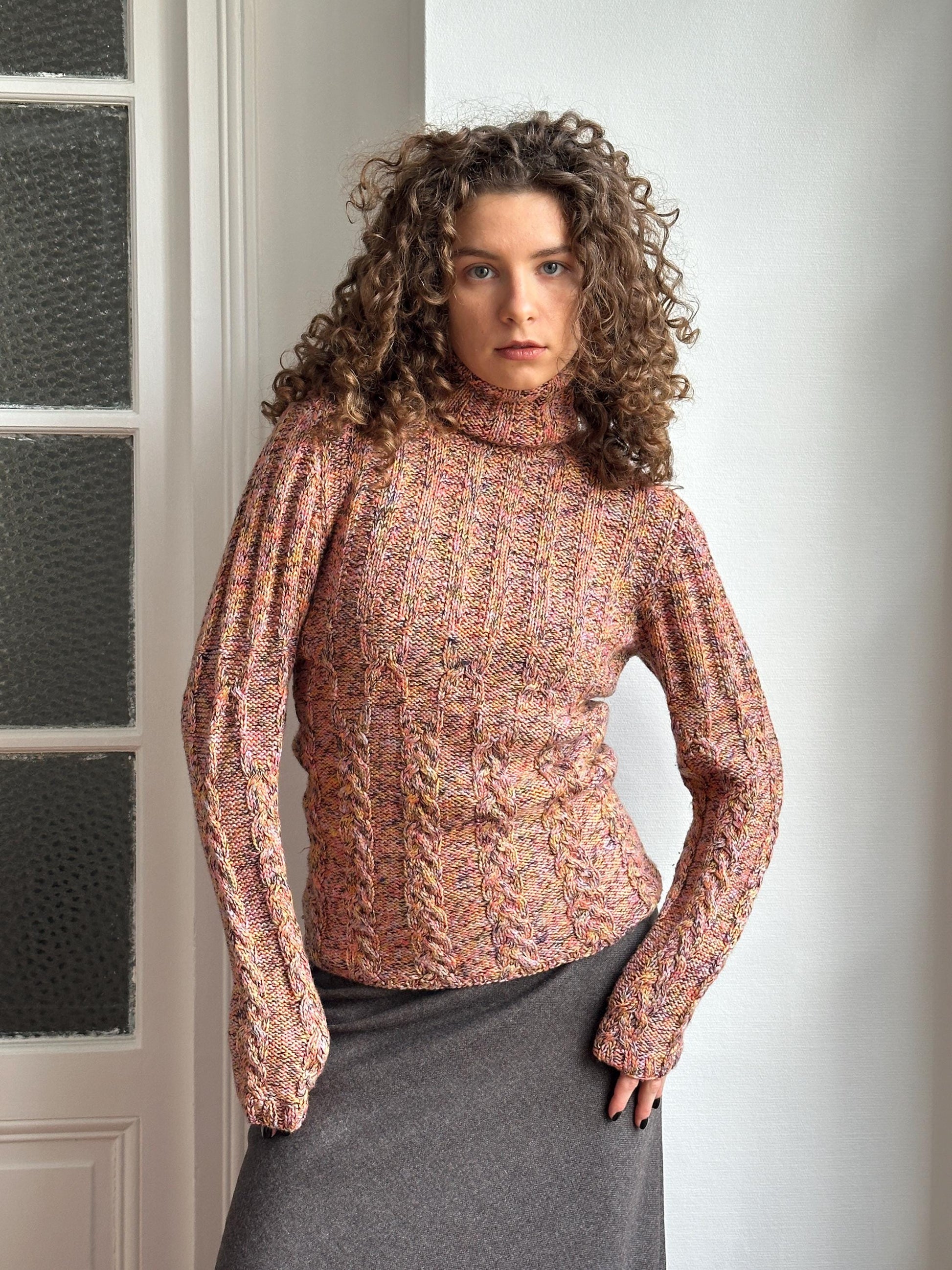 A woman with curly hair wearing a pink and orange knitted sweater is posing in a relaxed manner.