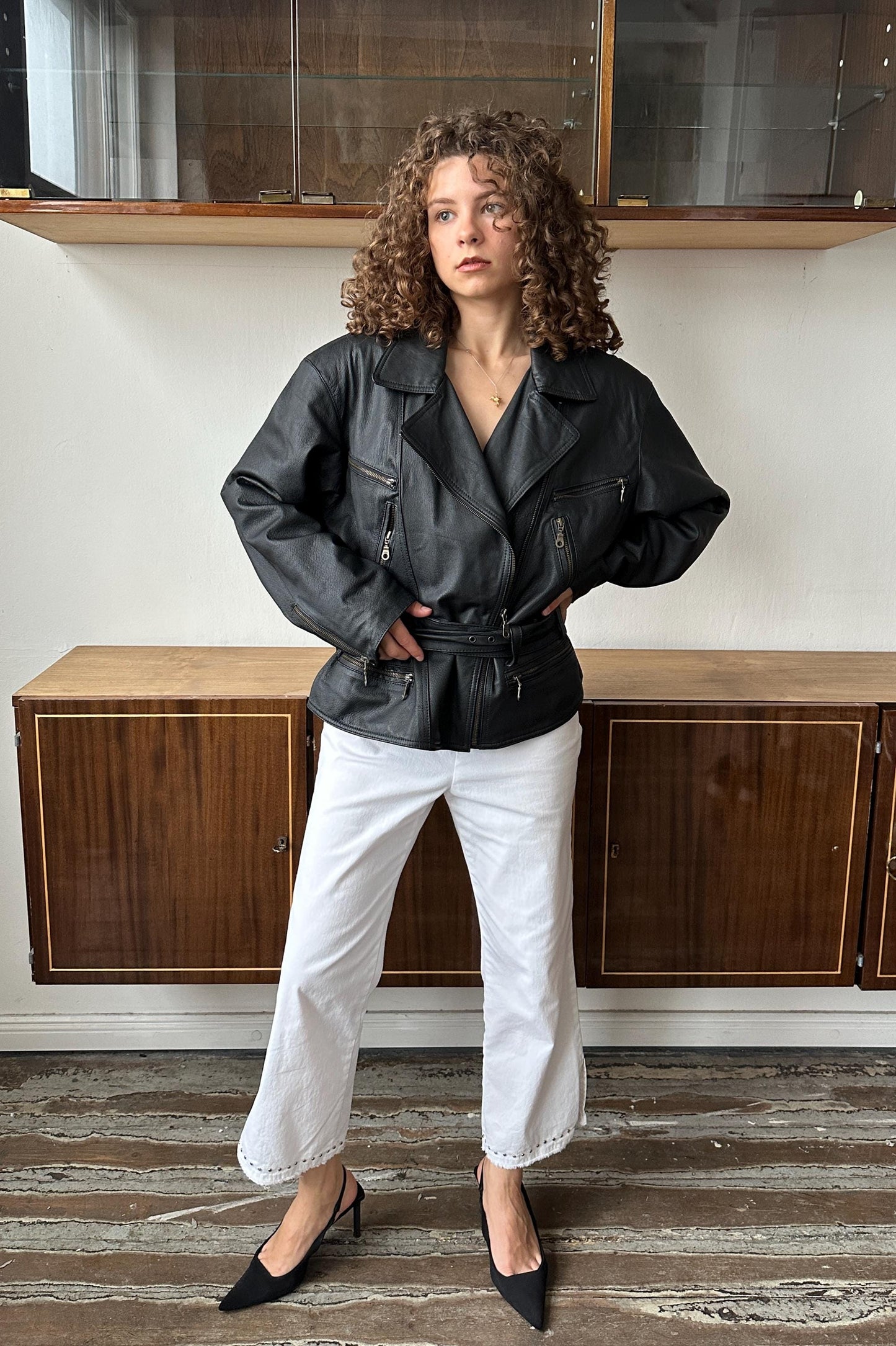 A woman with curly hair wearing a black leather jacket and white pants stands in front of a wooden cabinet.