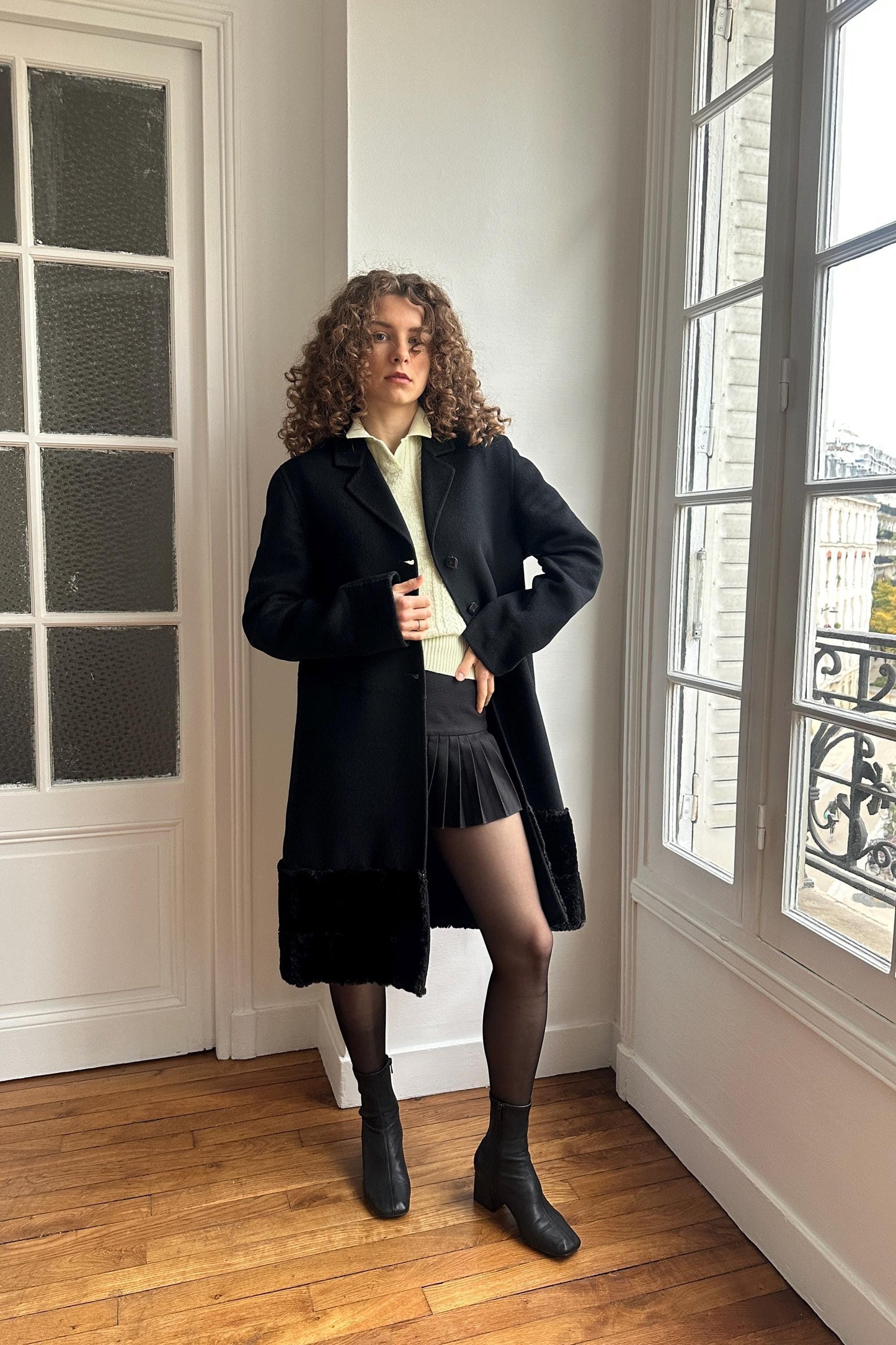 A woman with curly hair stands in a room, wearing a black coat, skirt, and boots.