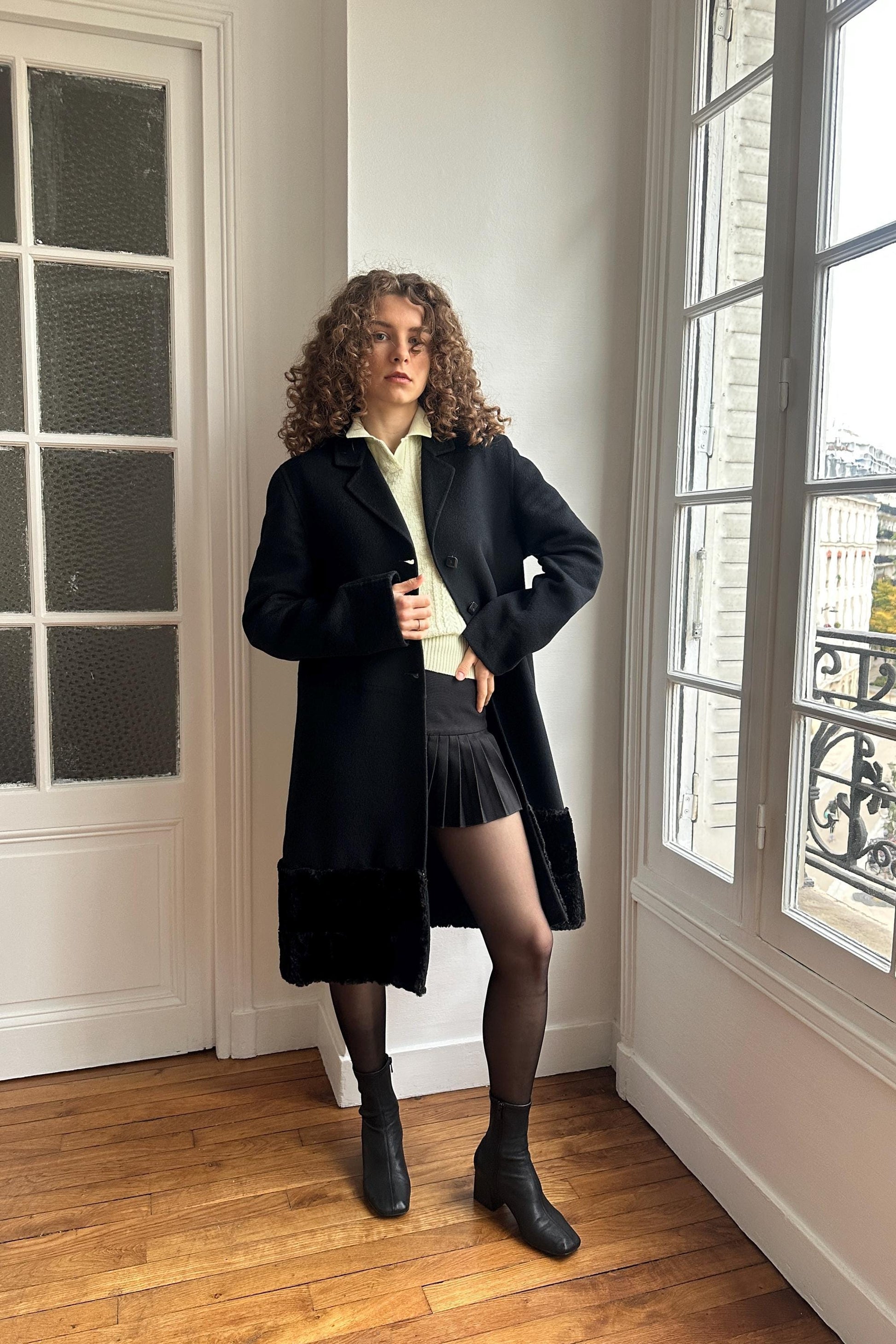 A woman with curly hair stands in a room, wearing a black coat, skirt, and boots.