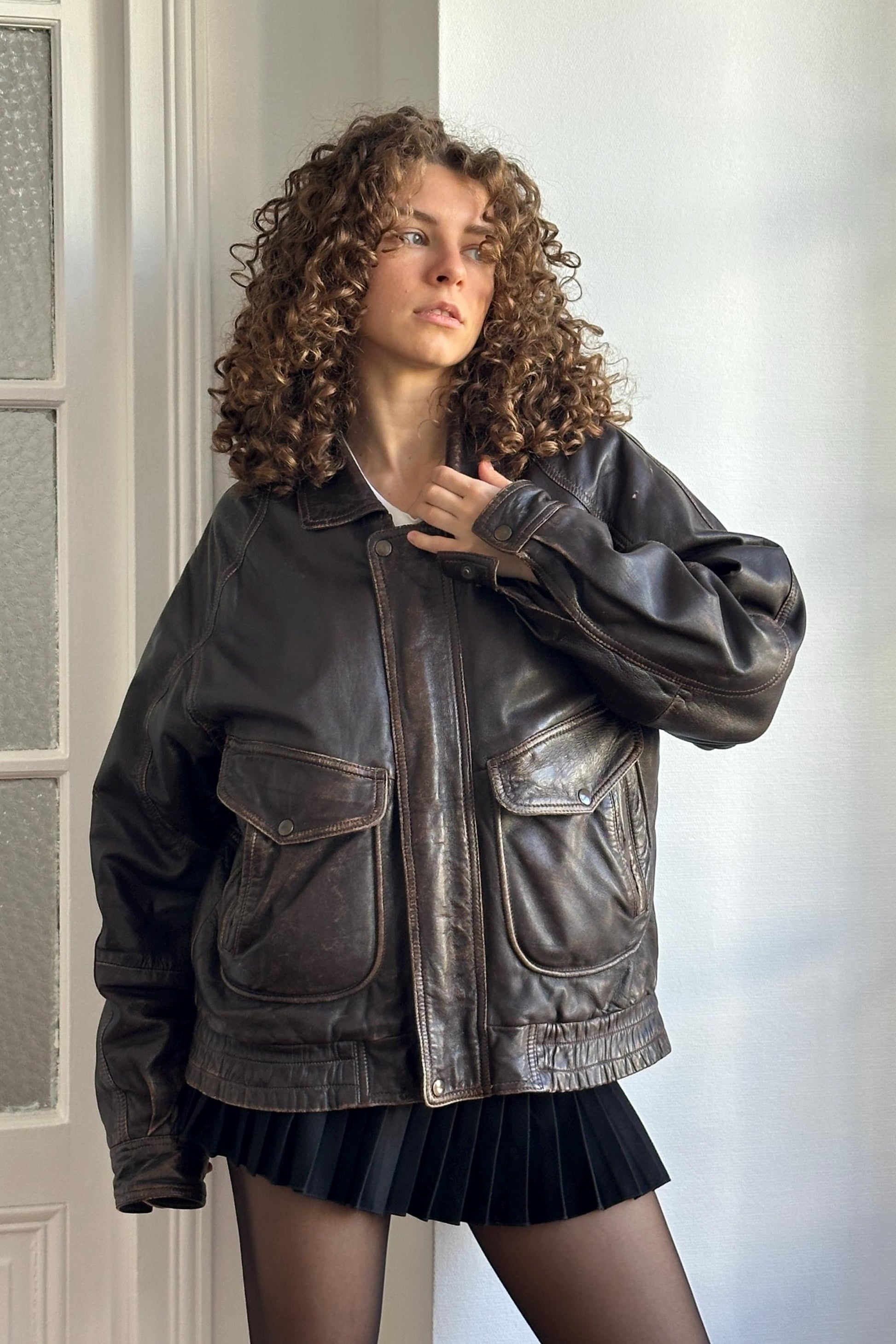A woman with curly hair wearing a brown leather jacket and a black skirt stands in front of a white door.