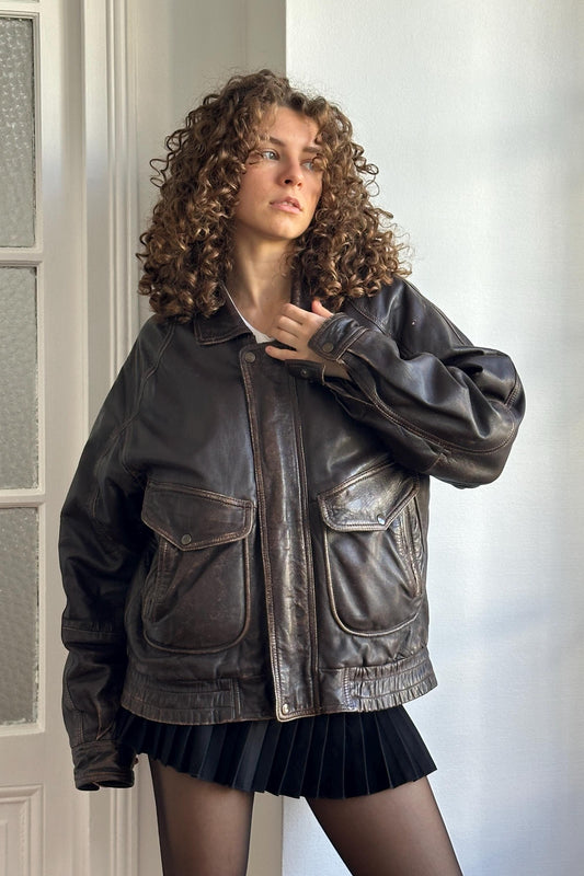 A woman with curly hair wearing a brown leather jacket and a black skirt stands in front of a white door.