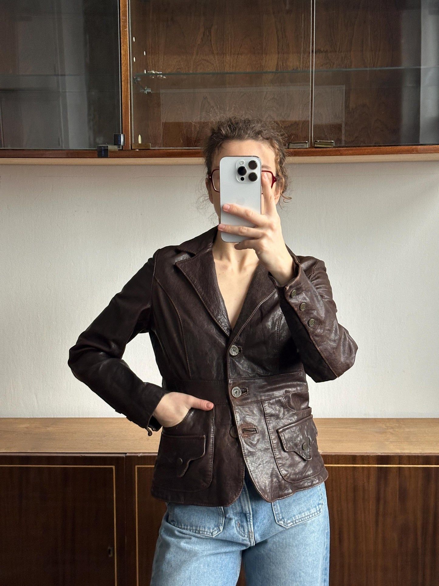 A person is taking a selfie with a smartphone, wearing a brown leather jacket and jeans.