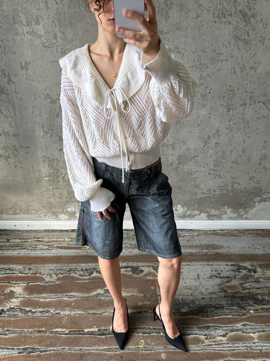 A person is taking a selfie with a white sweater, blue shorts, and black heels, standing in front of a concrete wall.