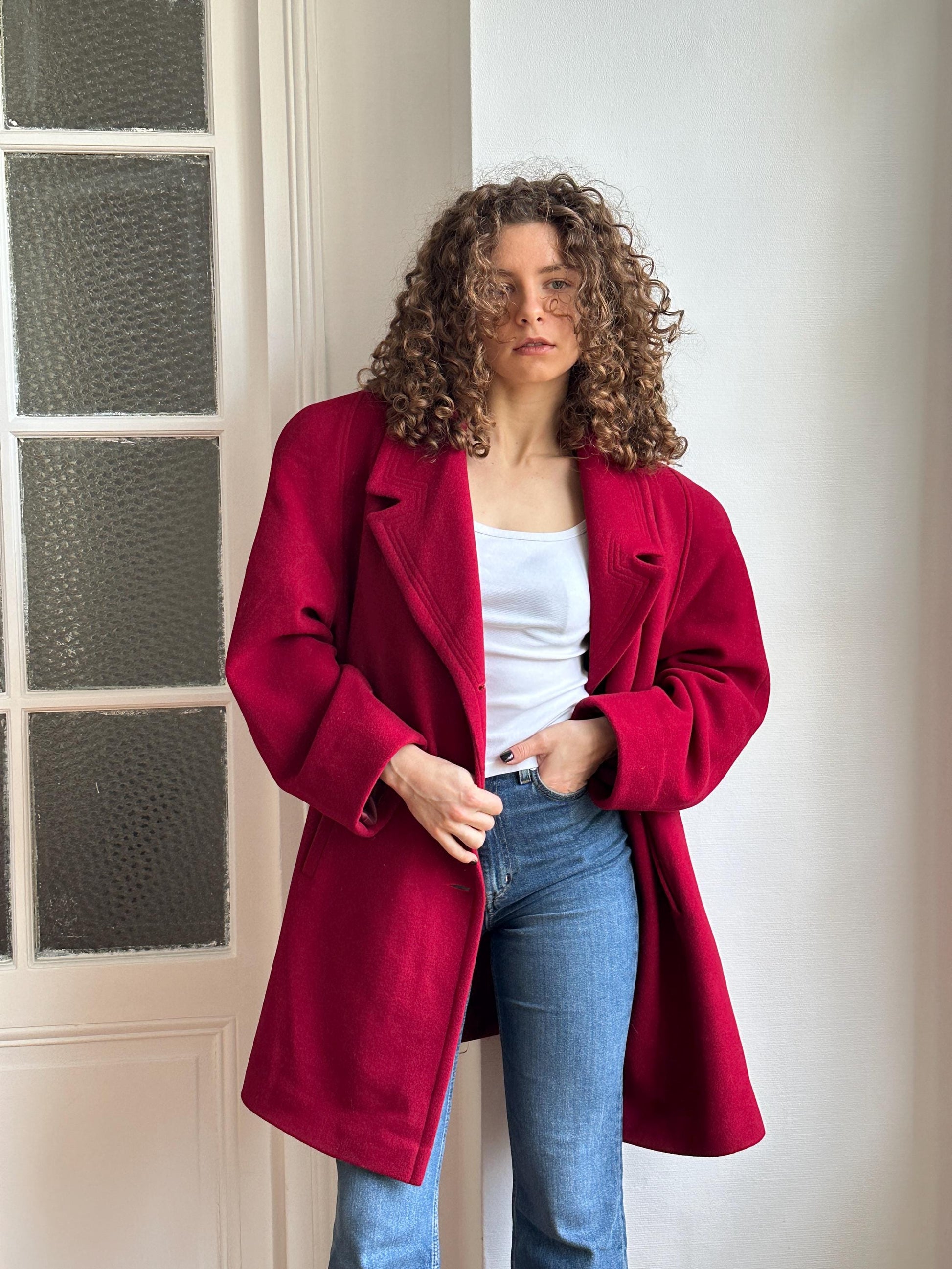 A woman with curly hair wearing a red coat is leaning against a white wall.