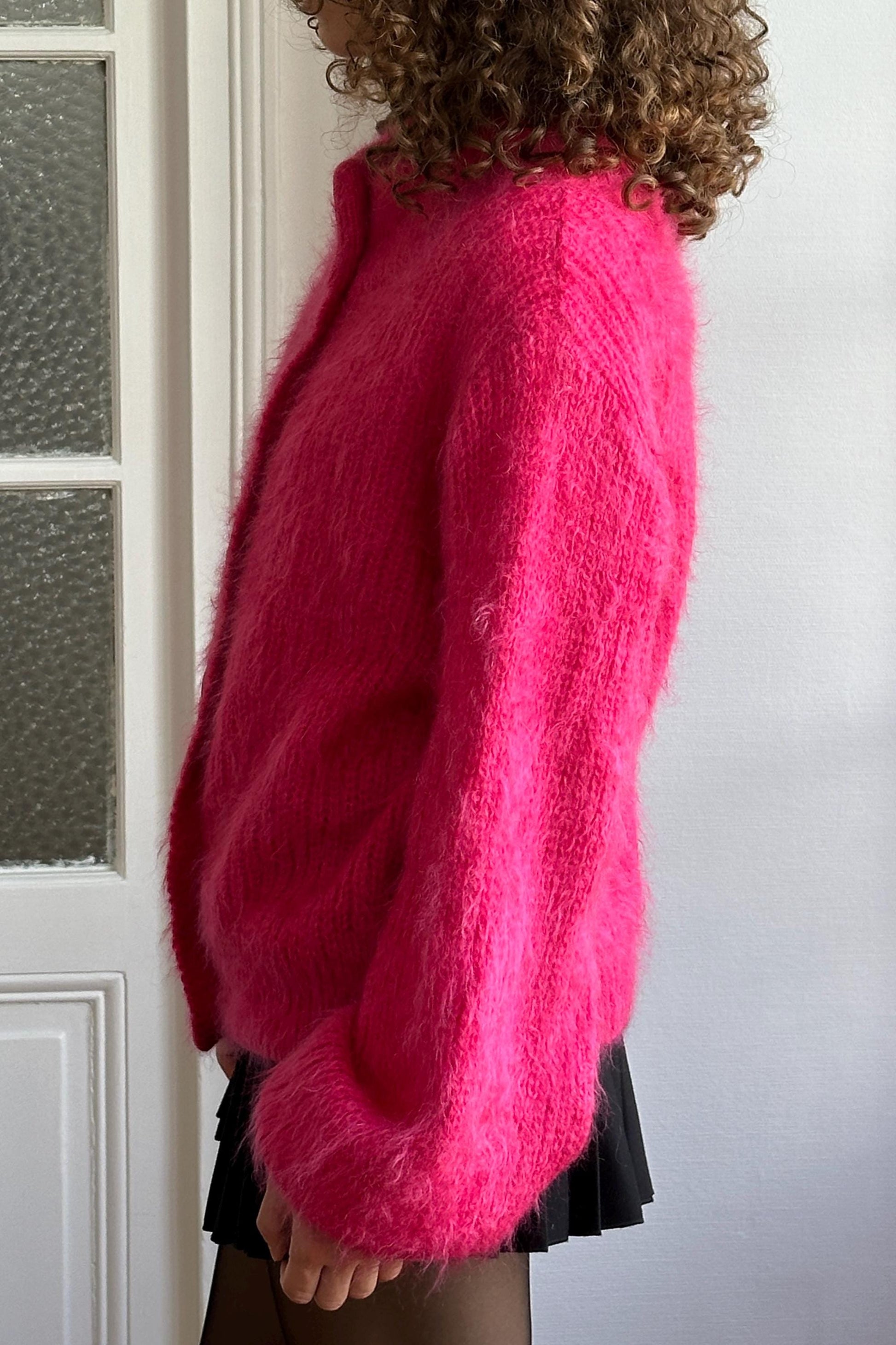 A person wearing a bright pink knitted cardigan stands in front of a white door.