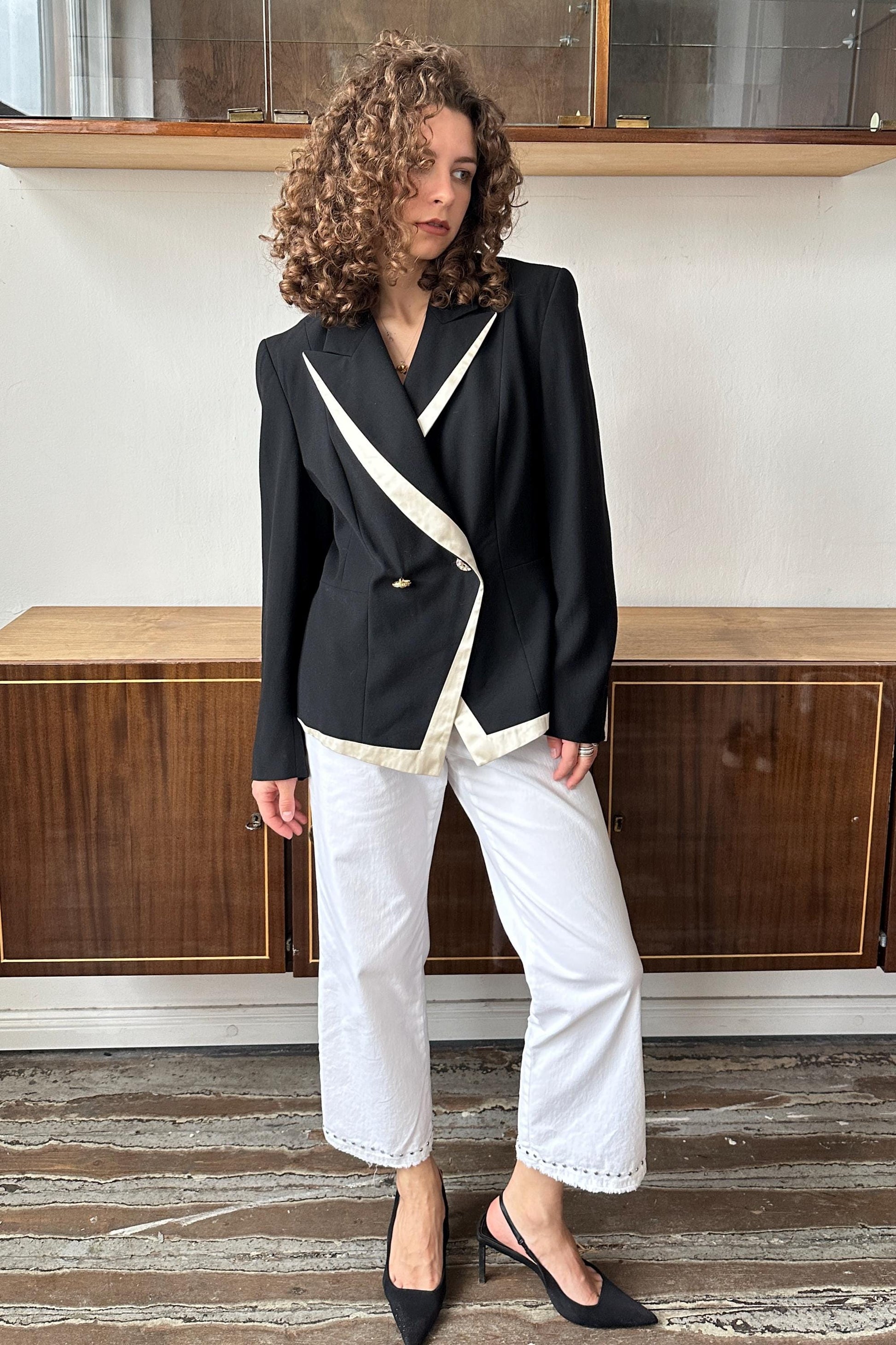A woman with curly hair wearing a black blazer and white pants stands in front of a wooden cabinet.