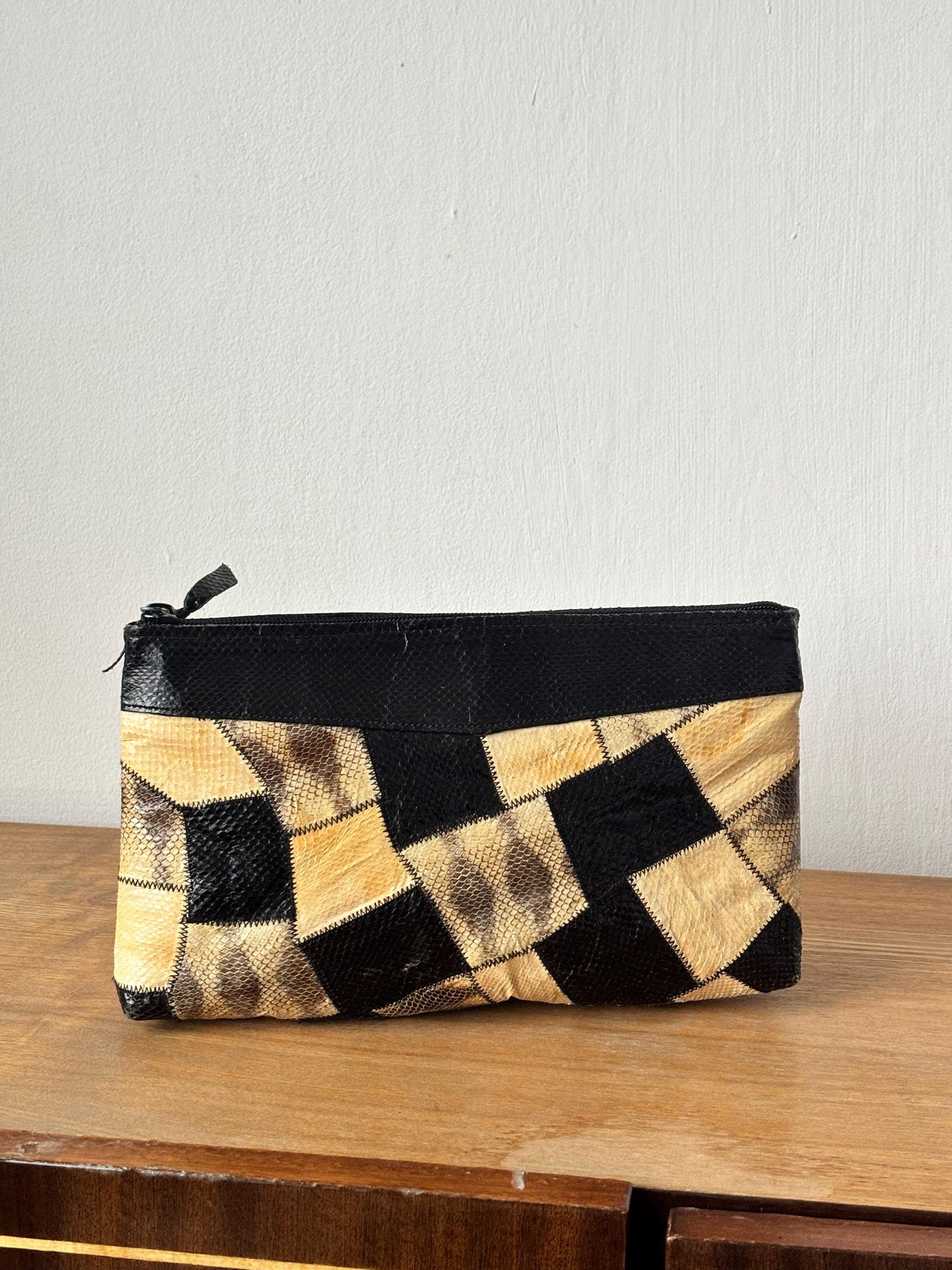 A black and tan patterned clutch purse is displayed on a wooden surface against a white wall.