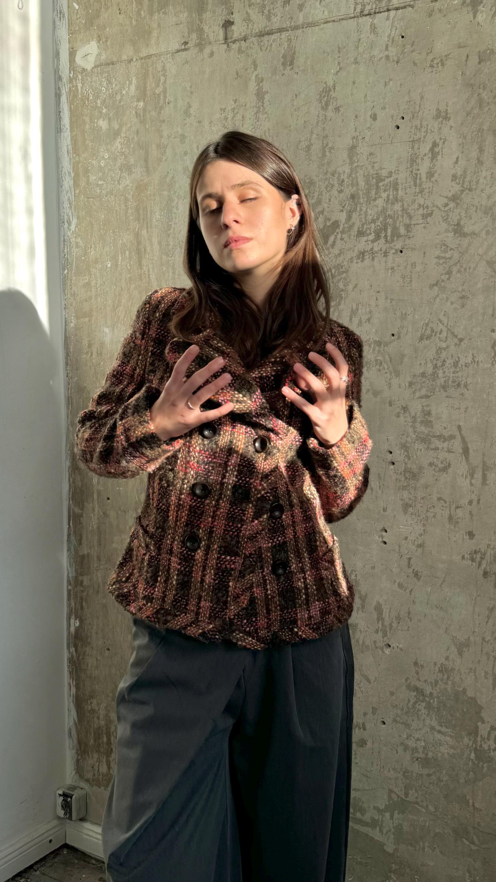 A woman with long brown hair stands in front of a concrete wall, her hands clasped together in a relaxed pose.