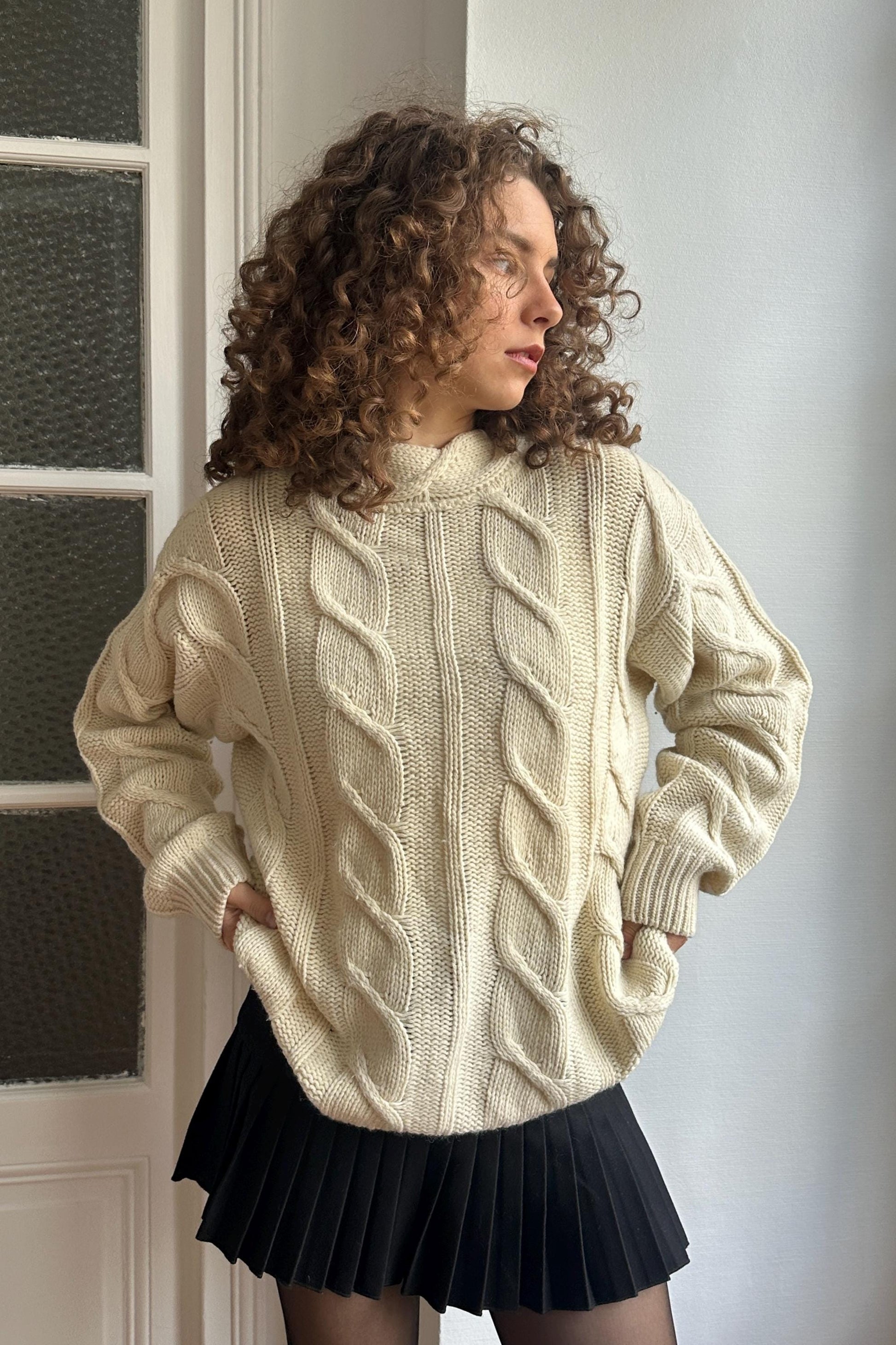 A woman with curly hair stands in front of a window, wearing a cream-colored sweater with a ribbed texture and a black pleated skirt.