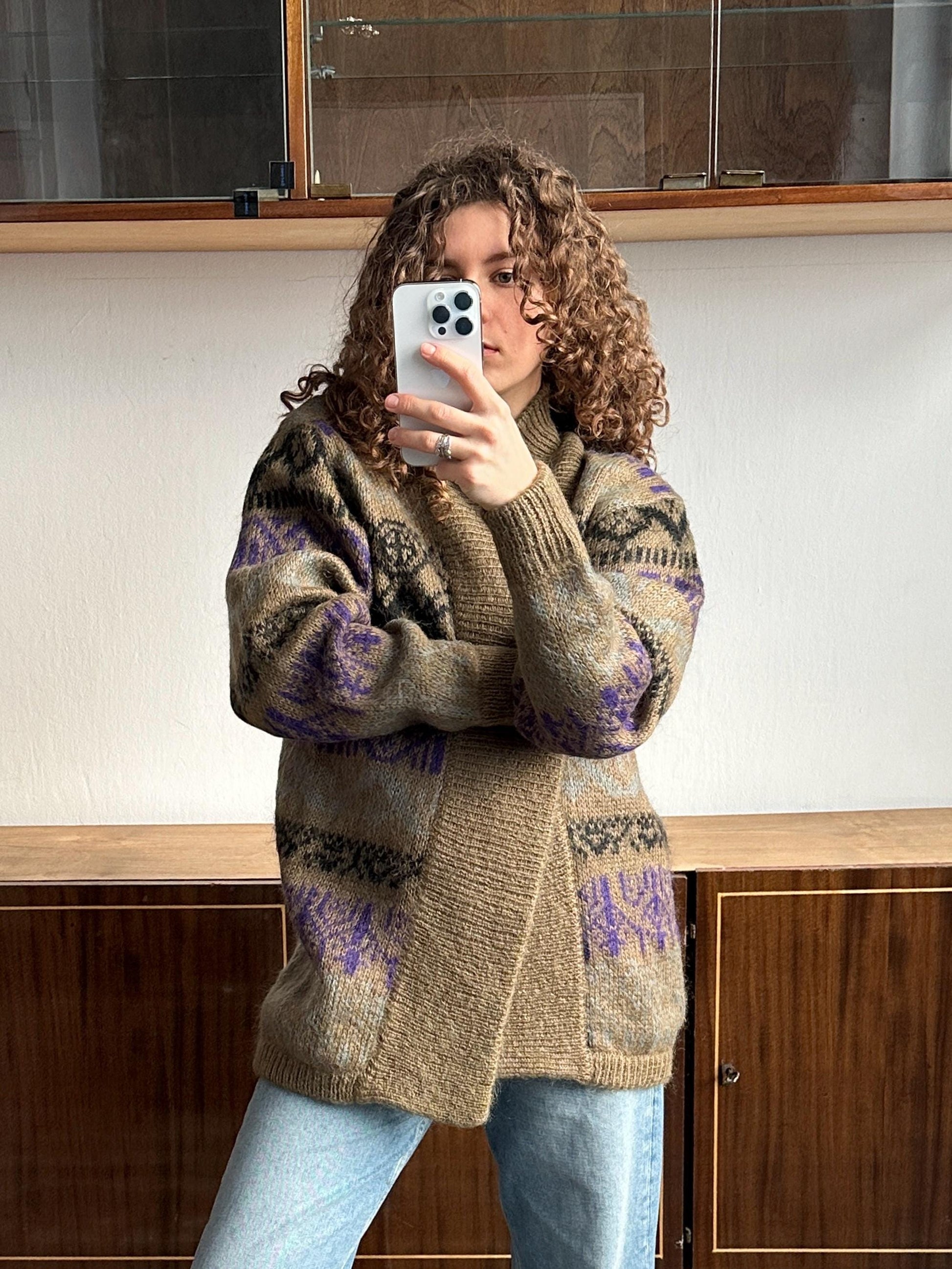 A woman with curly hair is taking a selfie with a white smartphone, wearing a patterned sweater and blue jeans.