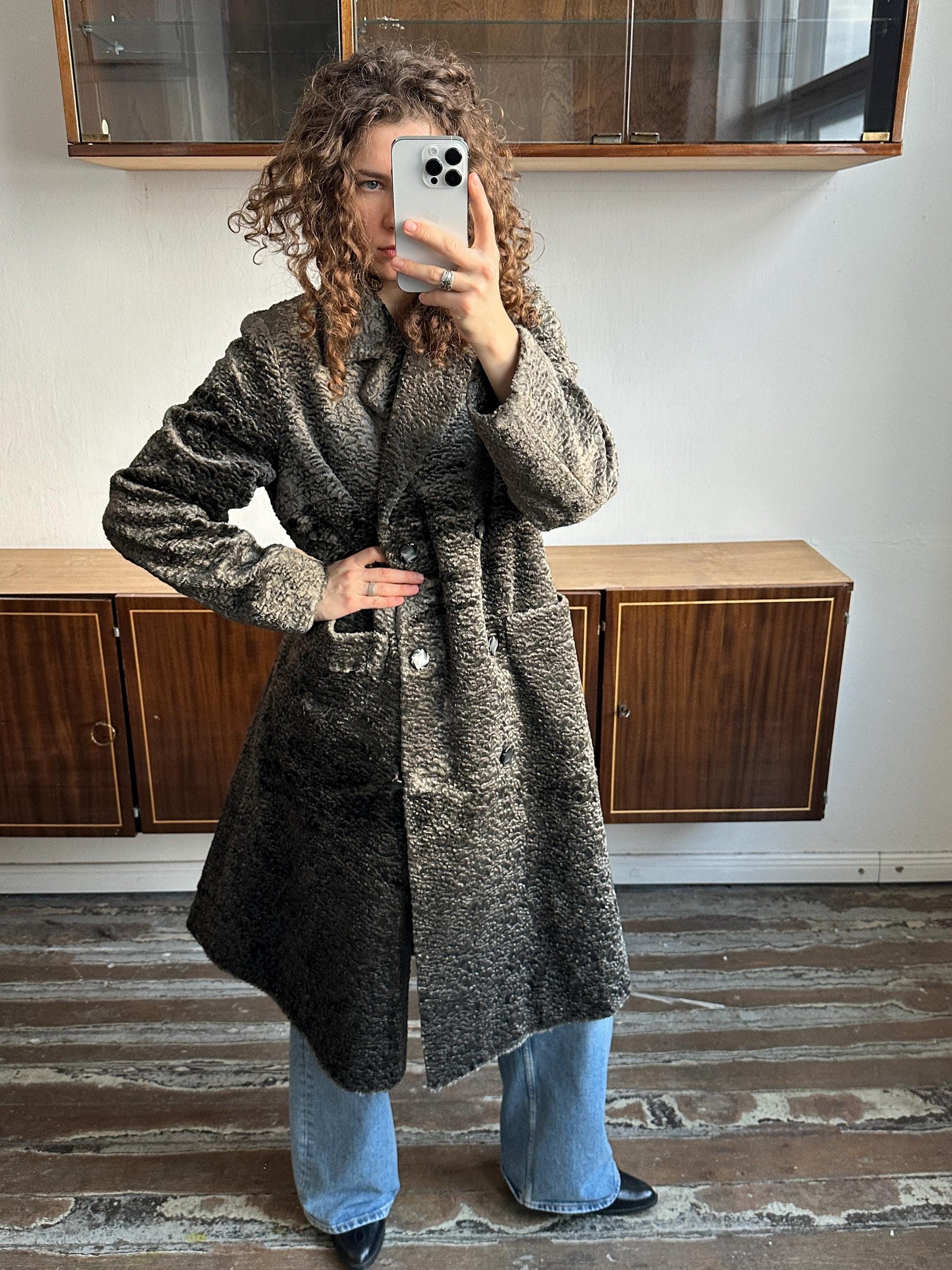 A woman with curly hair is taking a selfie with a smartphone, wearing a long gray coat and blue jeans.
