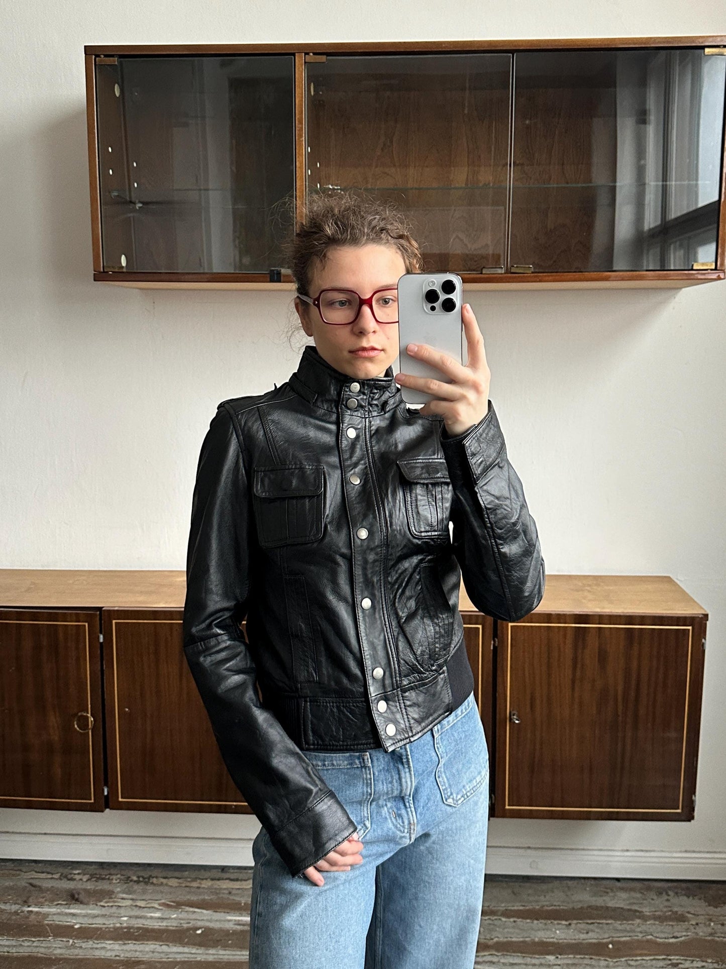 A person wearing a black leather jacket and jeans is taking a selfie with a smartphone in front of a wooden cabinet.
