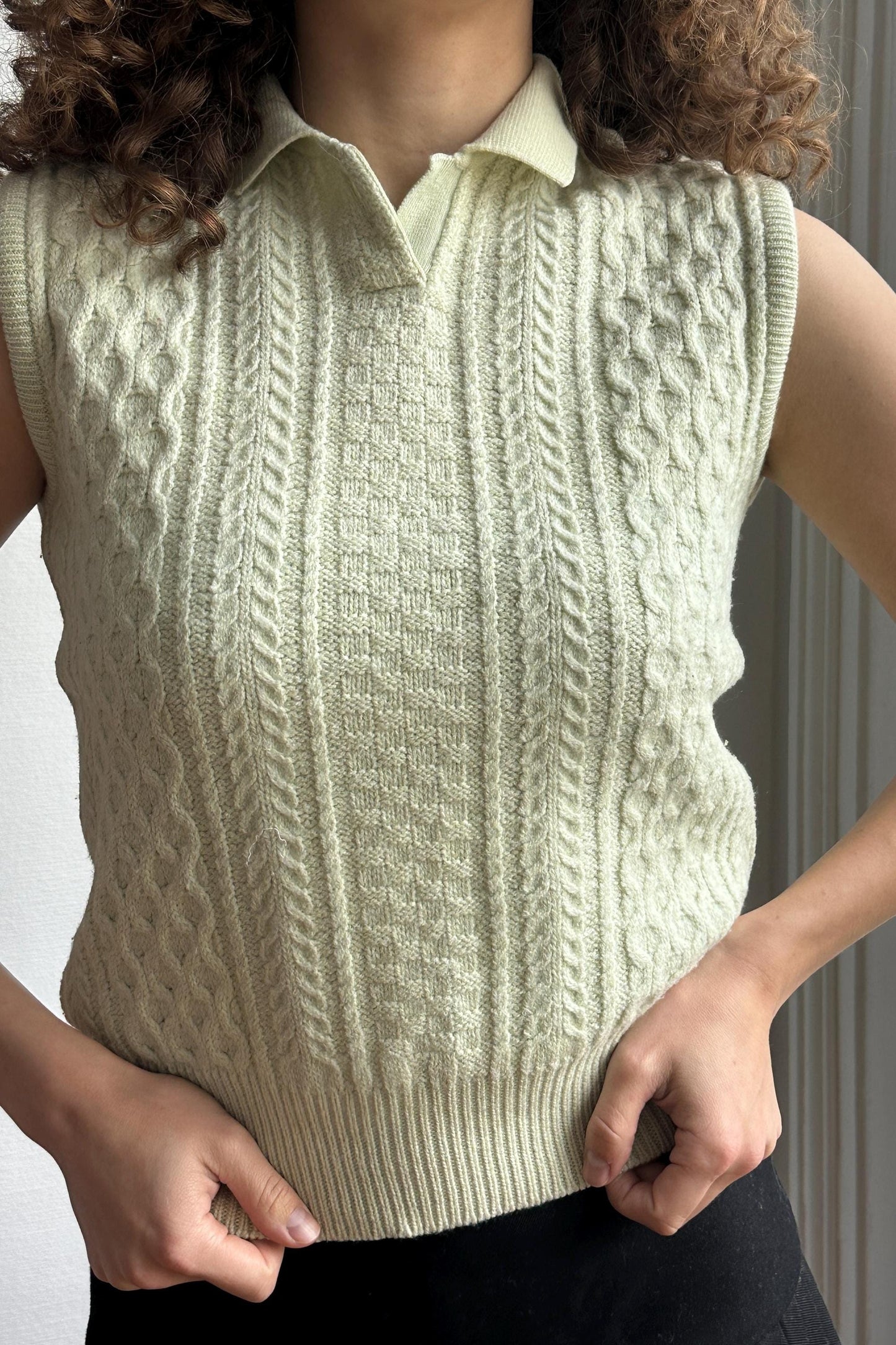 A person wearing a cream-colored knitted vest with a high collar and a patterned design.