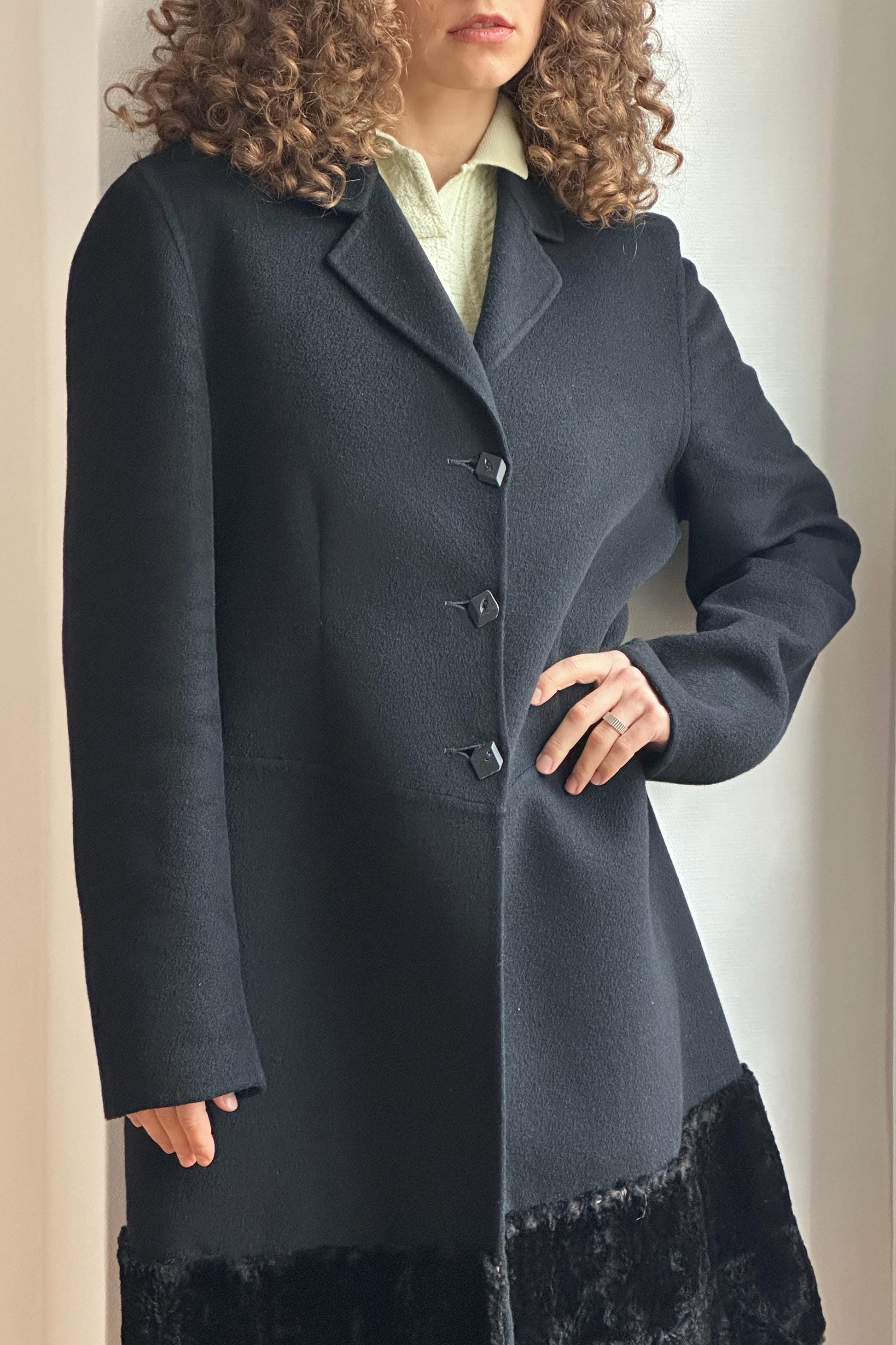 A woman with curly hair wearing a black coat with fur trim, standing in front of a white wall.