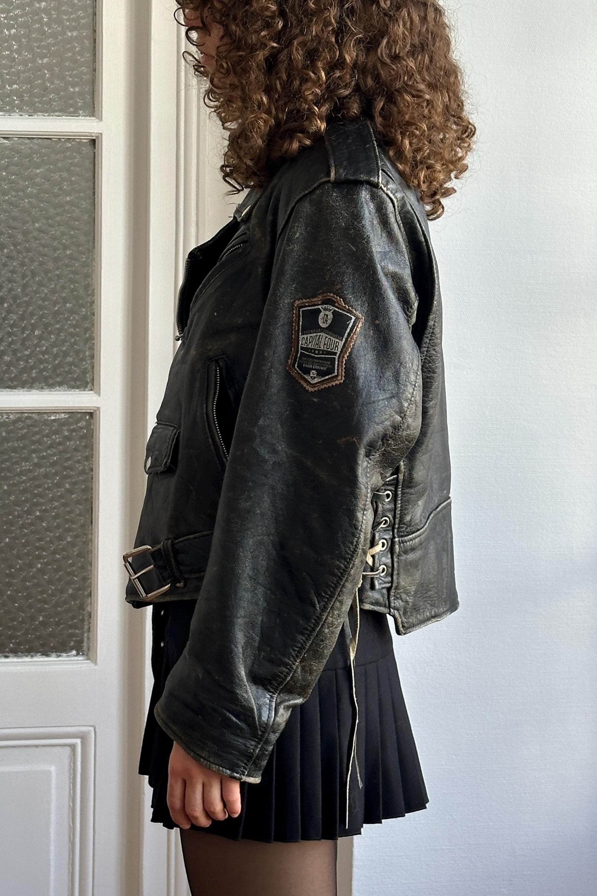 A person with curly hair wearing a black leather jacket and a black skirt stands in front of a white door.