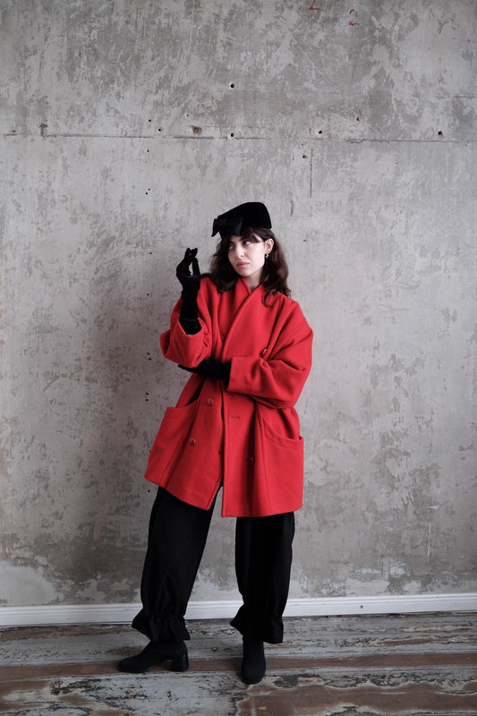 A person wearing a red coat and black pants stands in a room with a concrete wall.