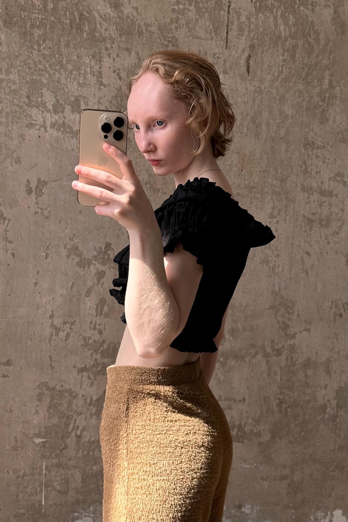 A woman is taking a selfie with her iPhone, wearing a black top and tan pants, against a textured concrete wall.