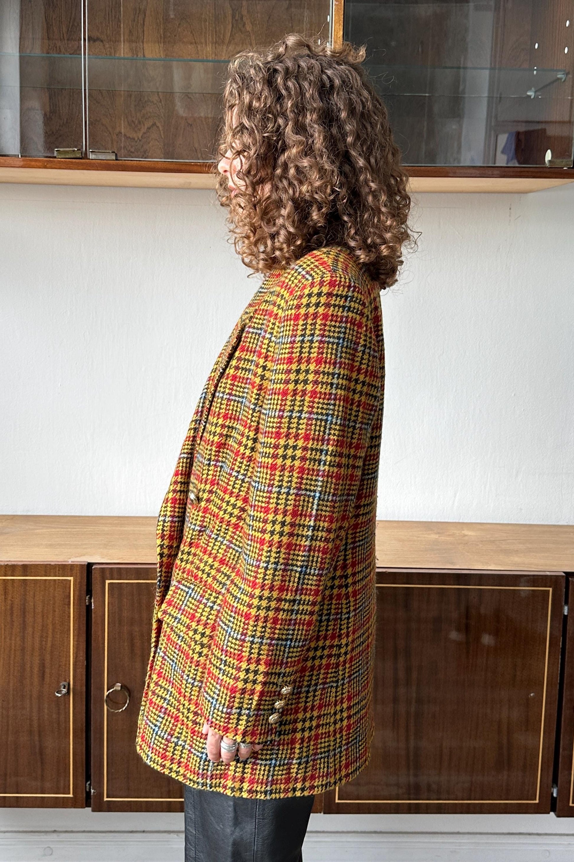 A person with curly hair wearing a colorful plaid jacket stands in front of a wooden cabinet.