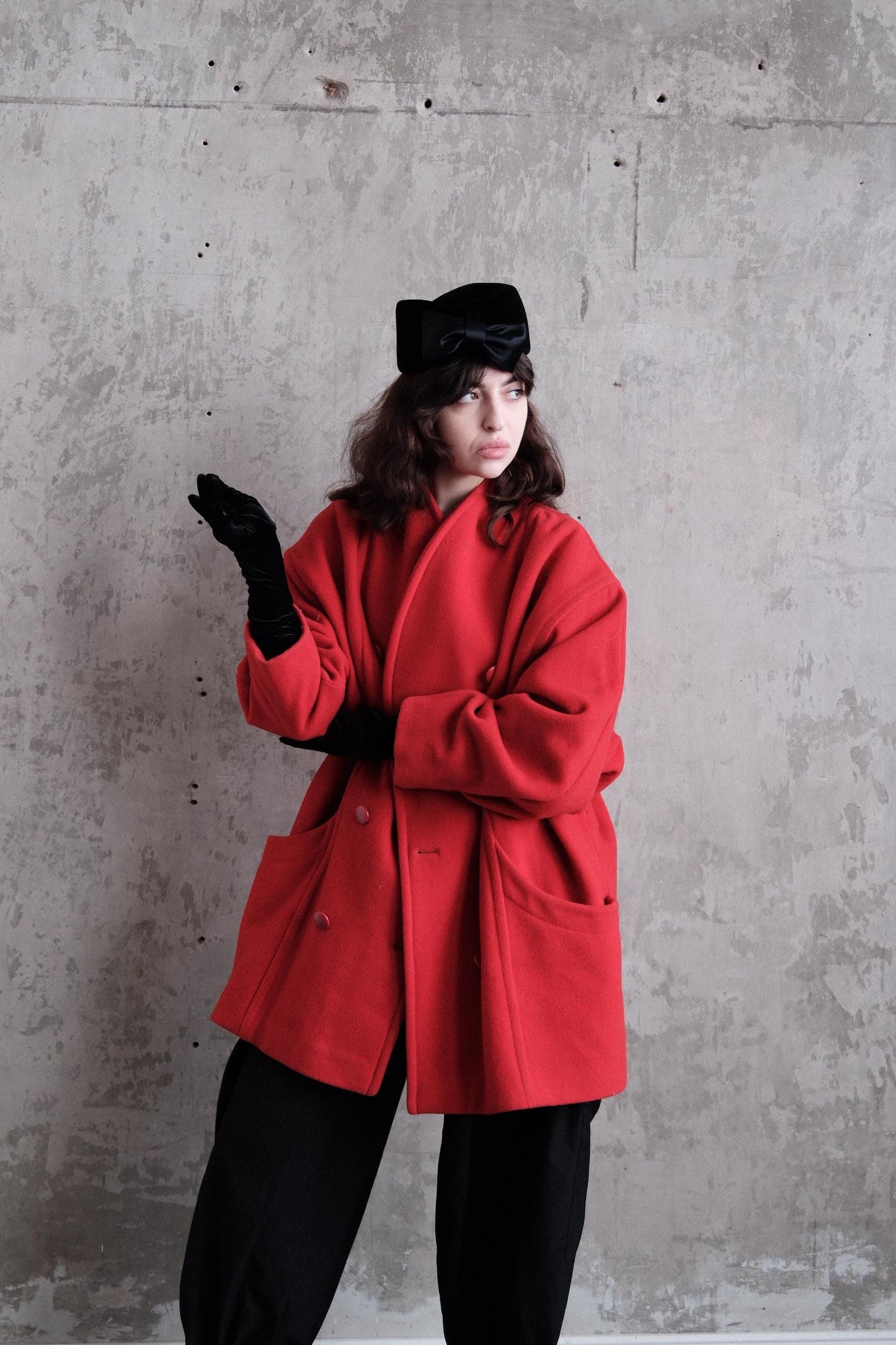 A person wearing a red coat and black gloves is standing against a concrete wall.