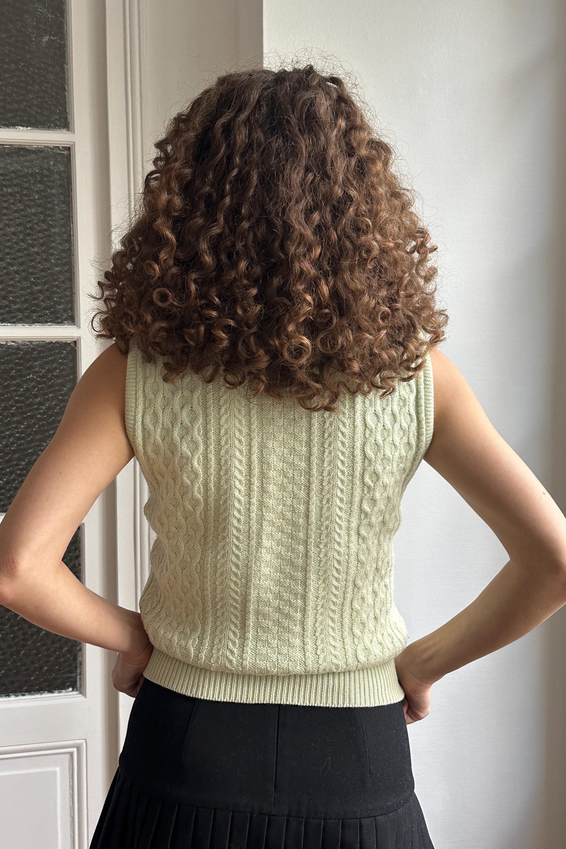 a person with curly hair wearing a white sleeveless sweater and a black skirt, standing in front of a white door.