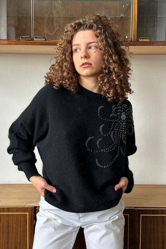 A young woman with curly hair wearing a black sweater with a floral design stands in front of a wooden cabinet.