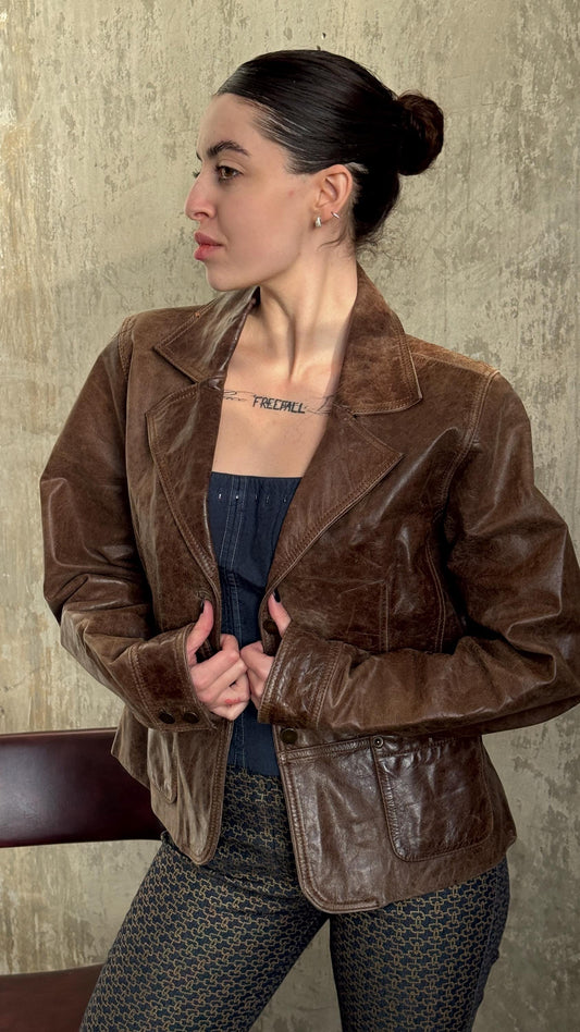 A woman wearing a brown leather jacket stands against a concrete wall, with her hair in a bun.
