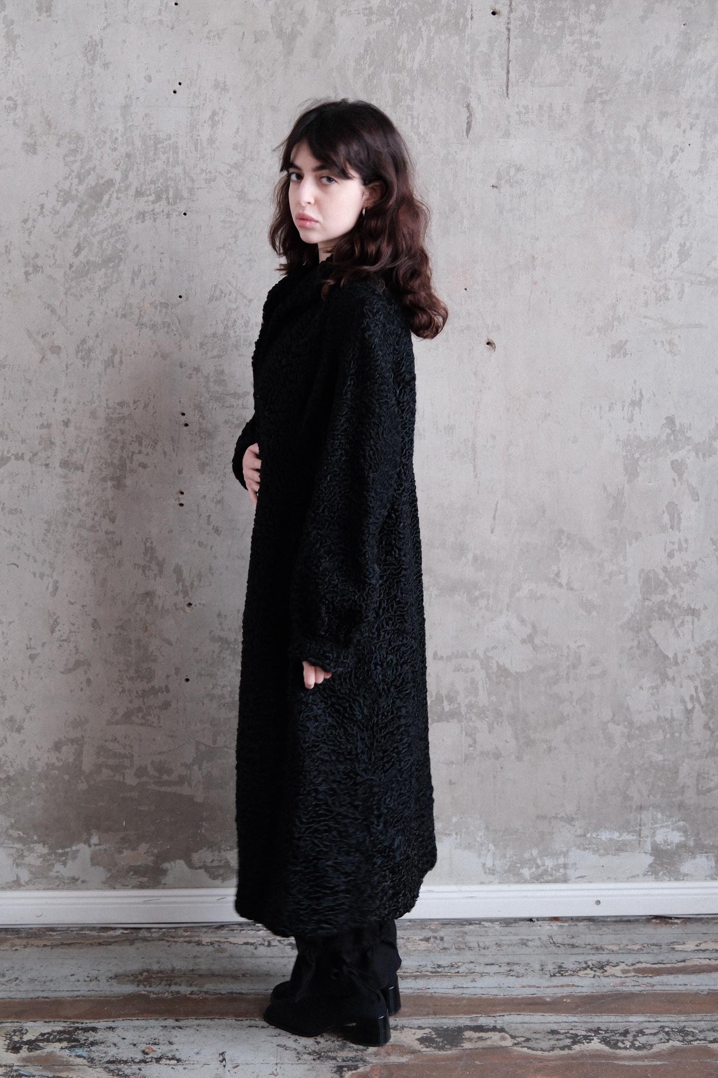 A woman in a long black coat stands against a concrete wall, her gaze directed off to the side.