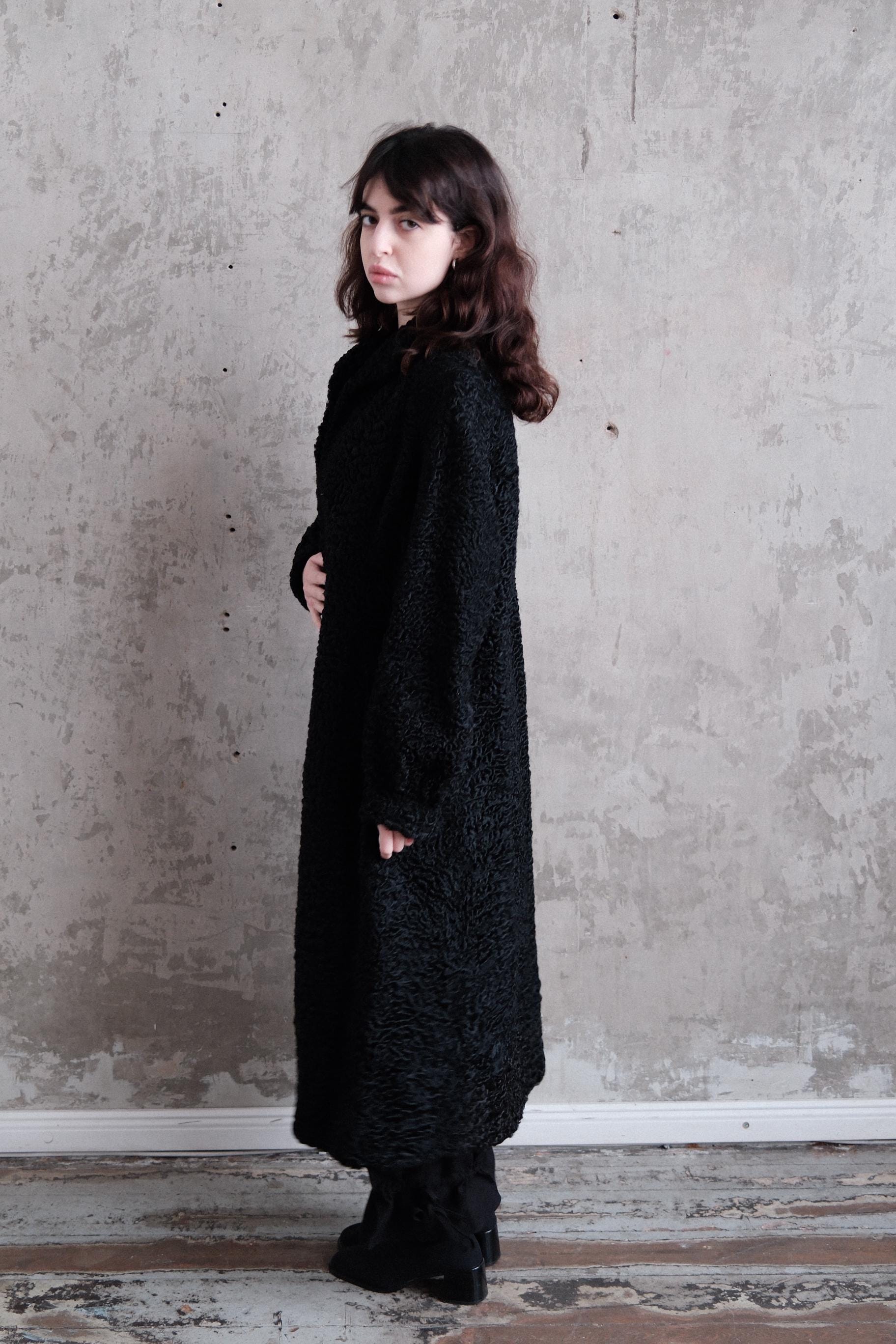 A woman in a long black coat stands against a concrete wall, her gaze directed off to the side.