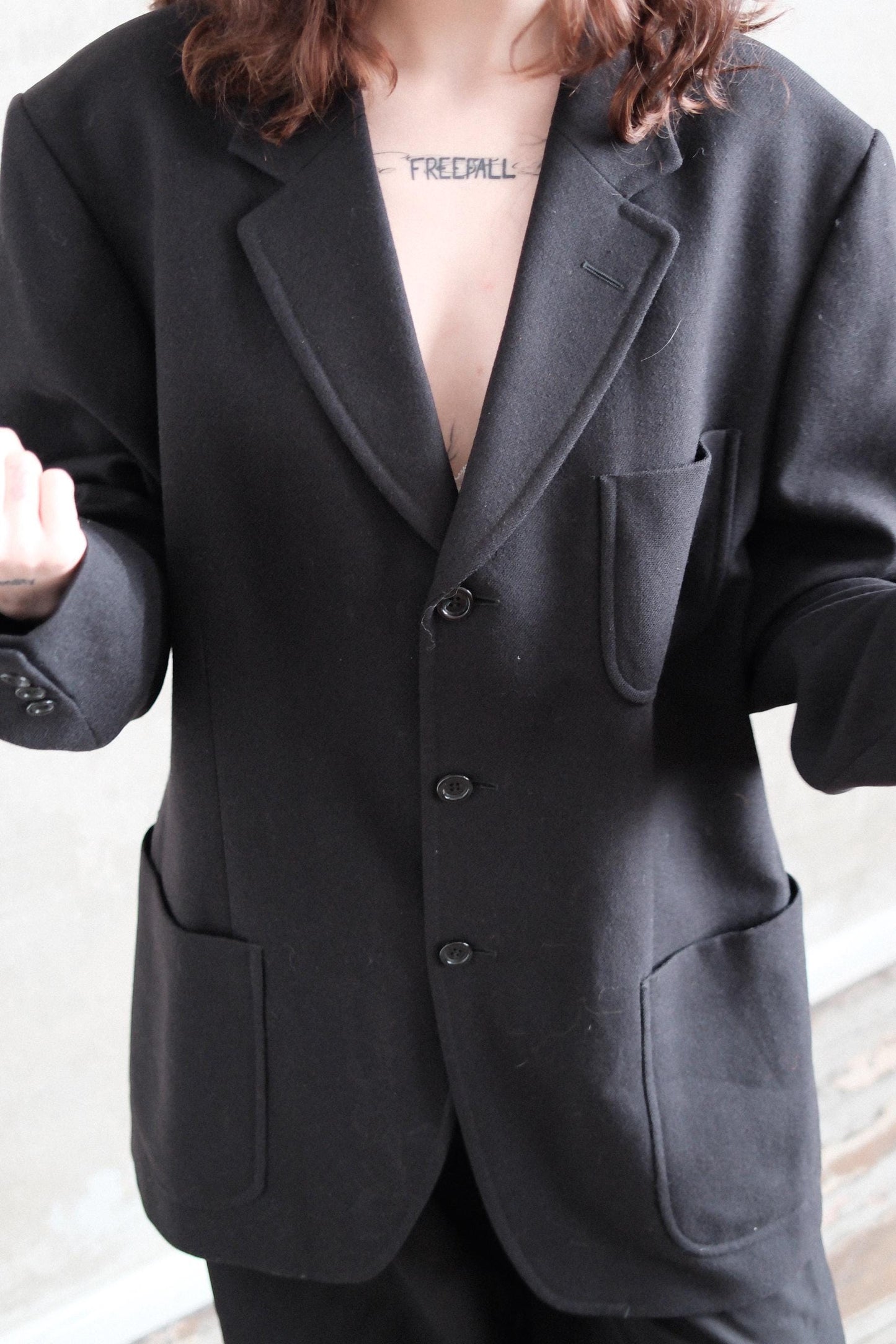 A person wearing a black blazer and standing in front of a white wall.