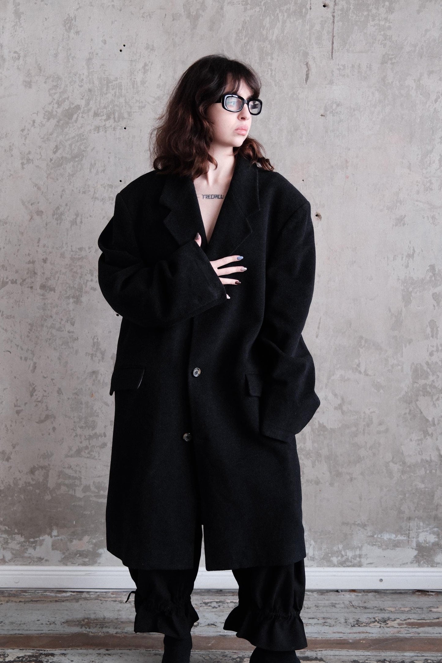 A person wearing a black coat and glasses stands against a plain gray wall.